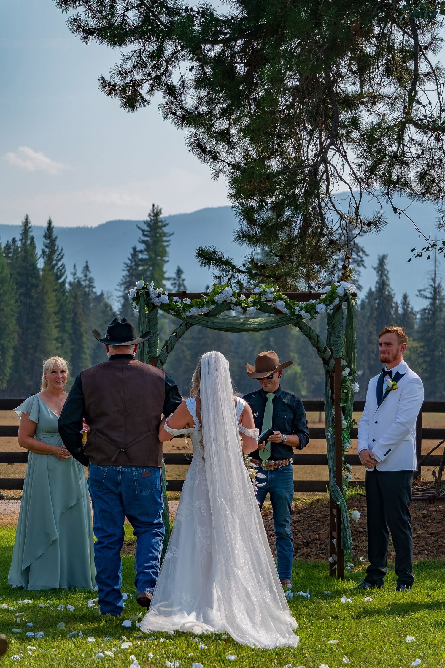 An outdoor wedding ceremony takes place under an arch with mountain and forest views.