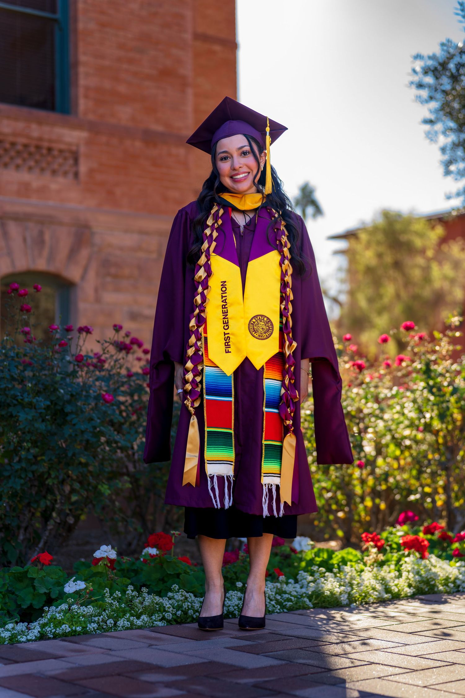 Graduate in purple commencement attire standing on stone pathway near blooming flowers.