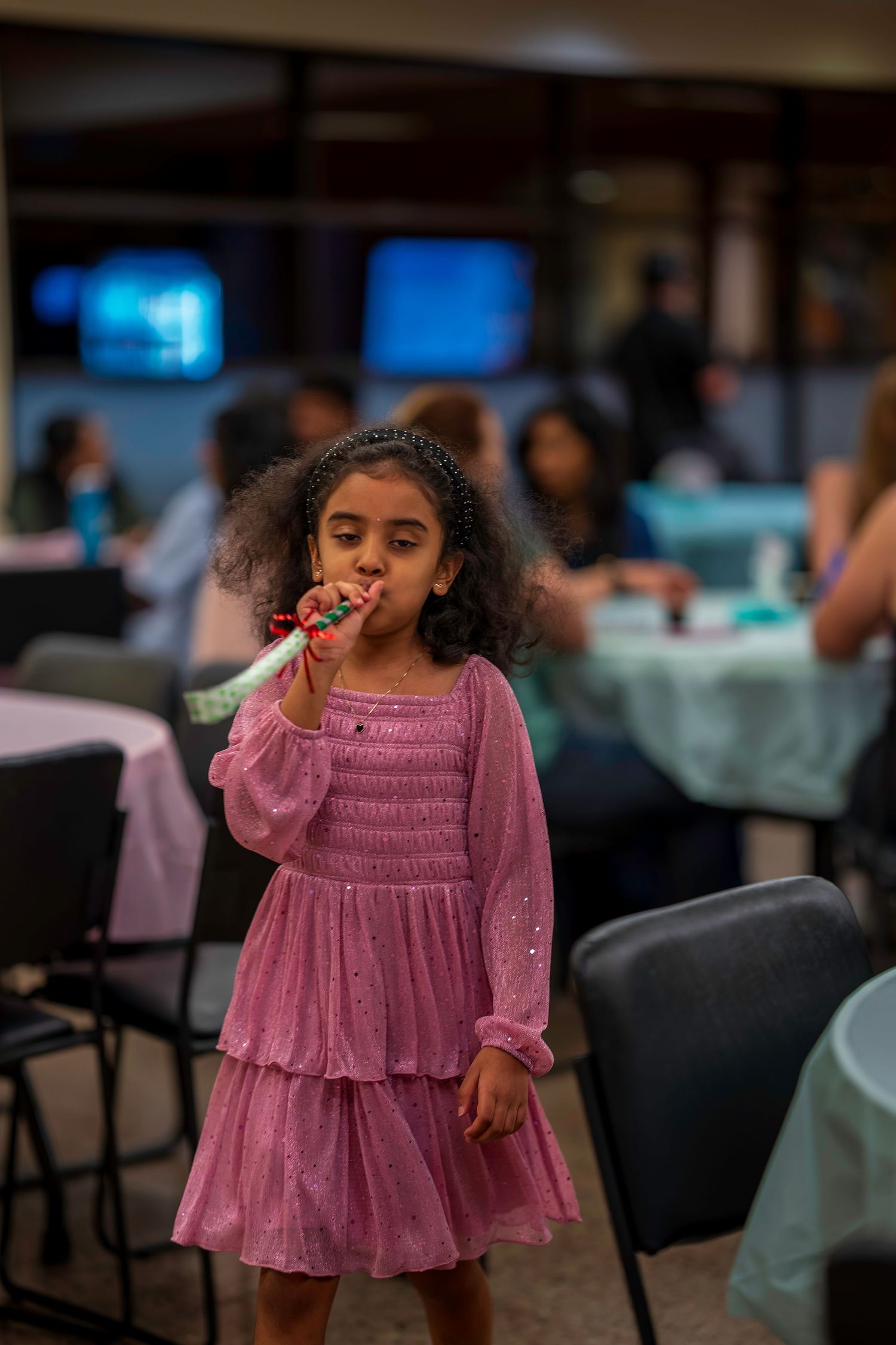 Young girl in pink dress blowing party horn at indoor celebration event.