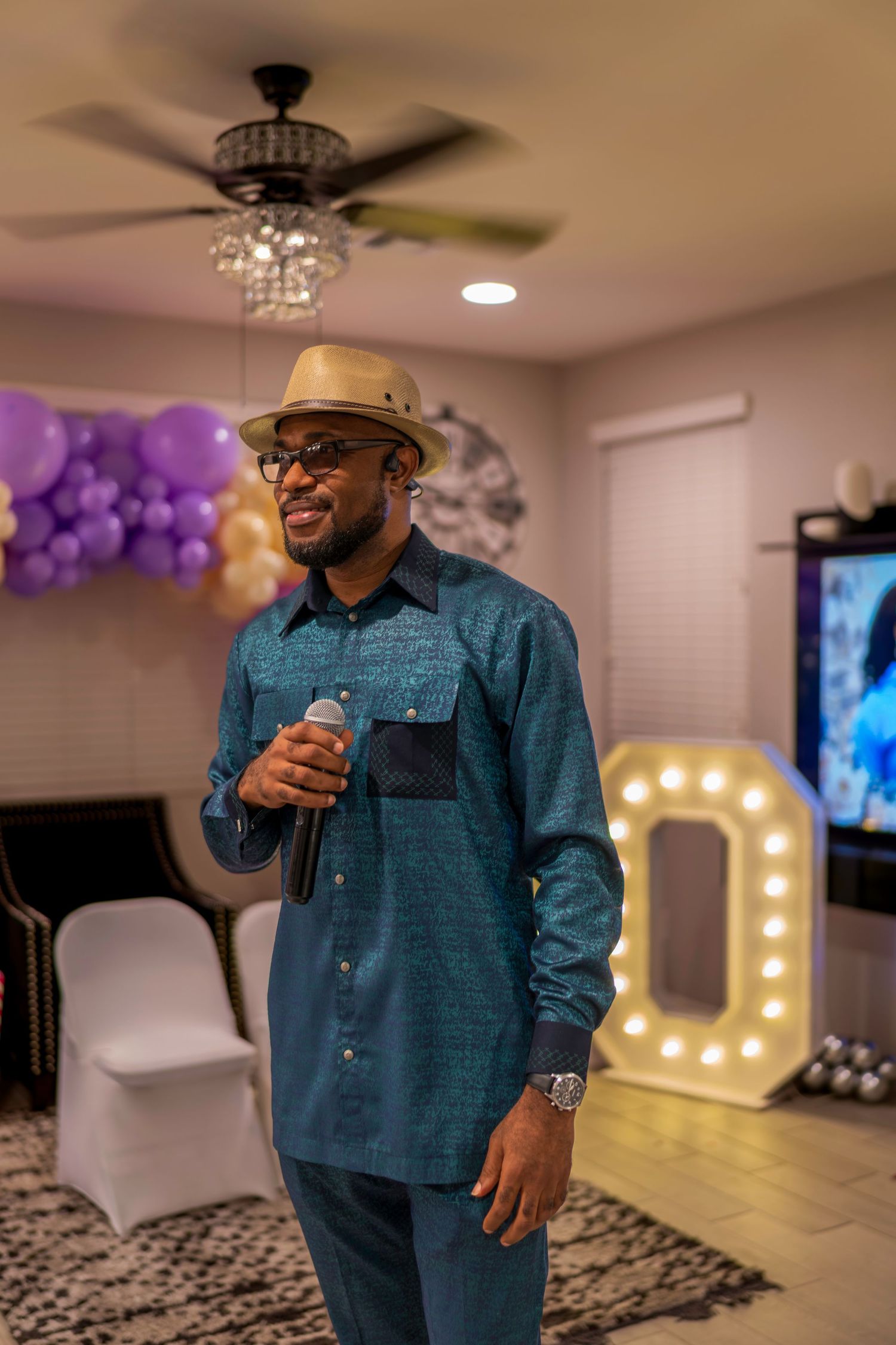 Black man in teal shirt and hat speaking into microphone at indoor celebration with balloons.