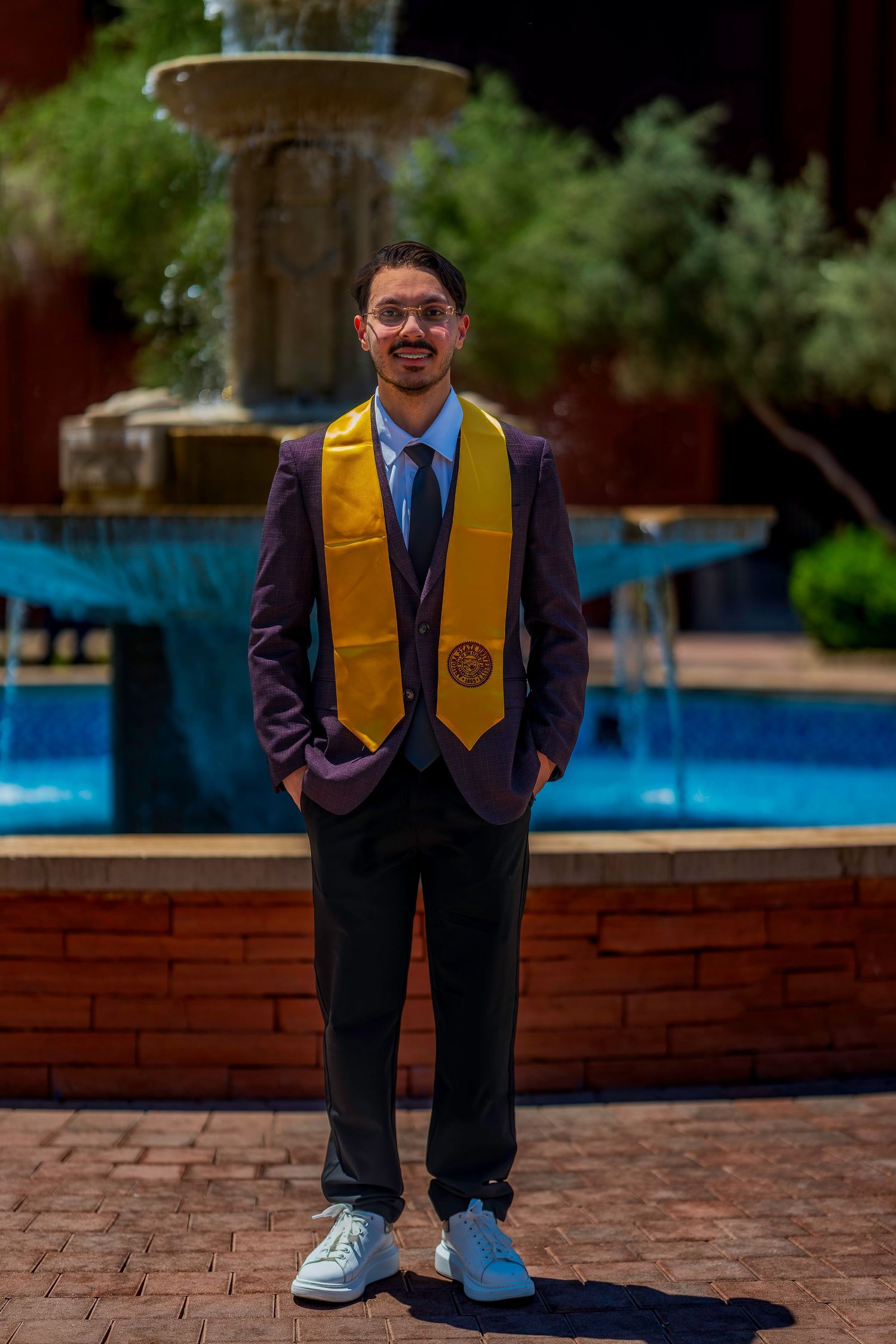 Young graduate in maroon suit and gold stole poses by fountain on sunny campus.