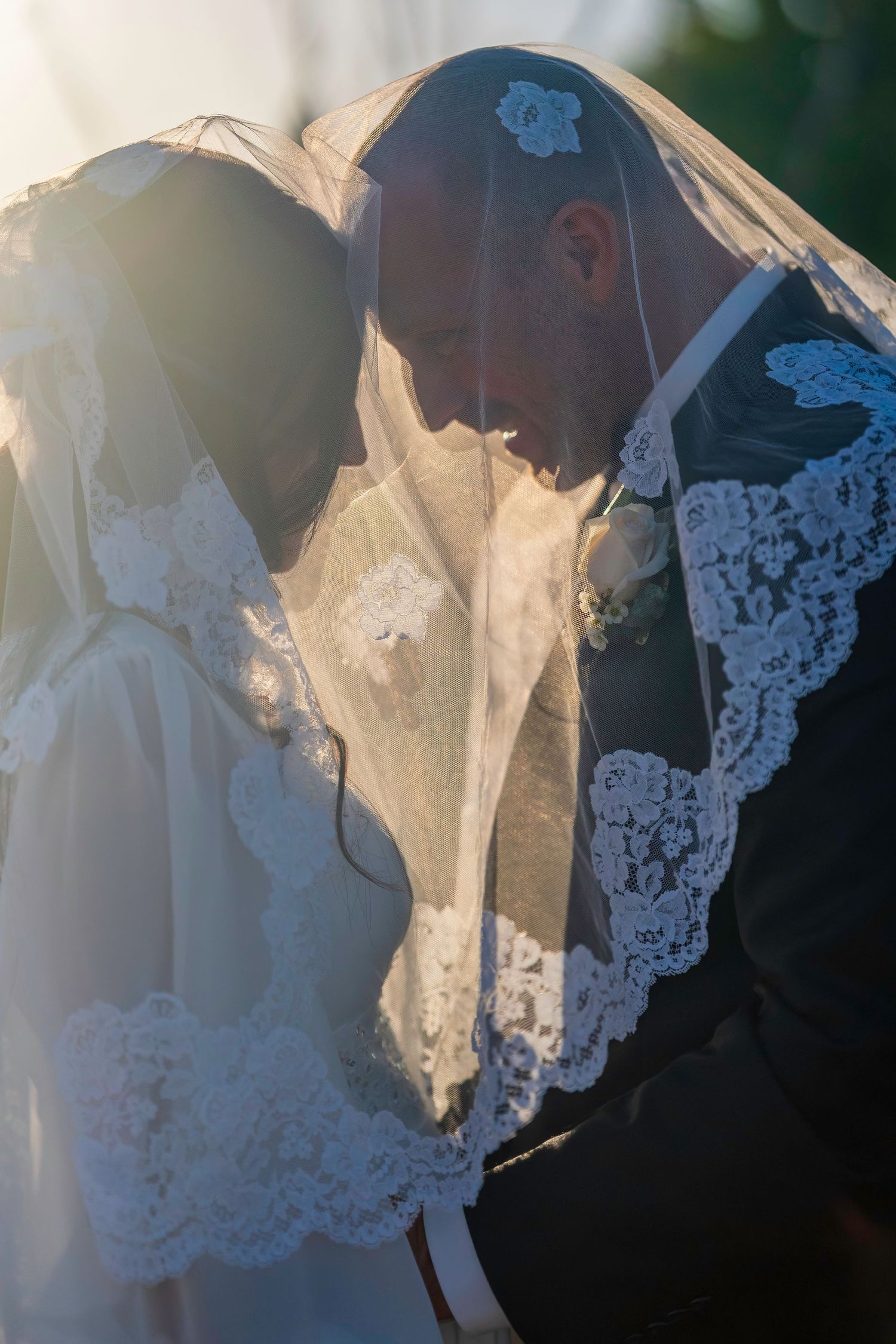 Romantic bride and groom sharing intimate moment under lace wedding veil with golden sunlight backdrop.