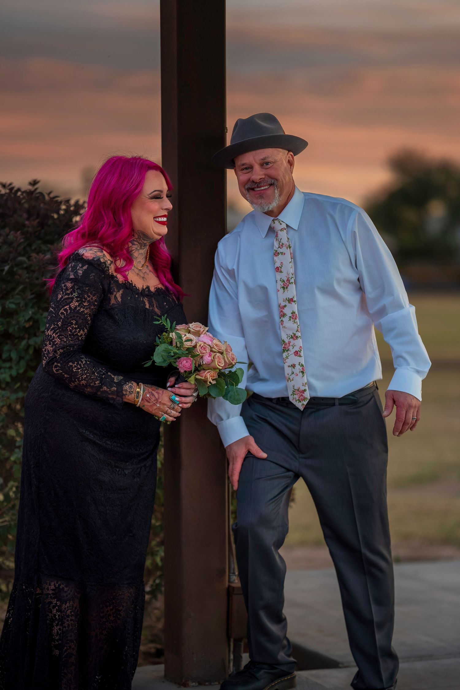Couple in wedding attire at sunset, woman with pink hair in black lace dress holding bouquet, man in hat and floral tie.