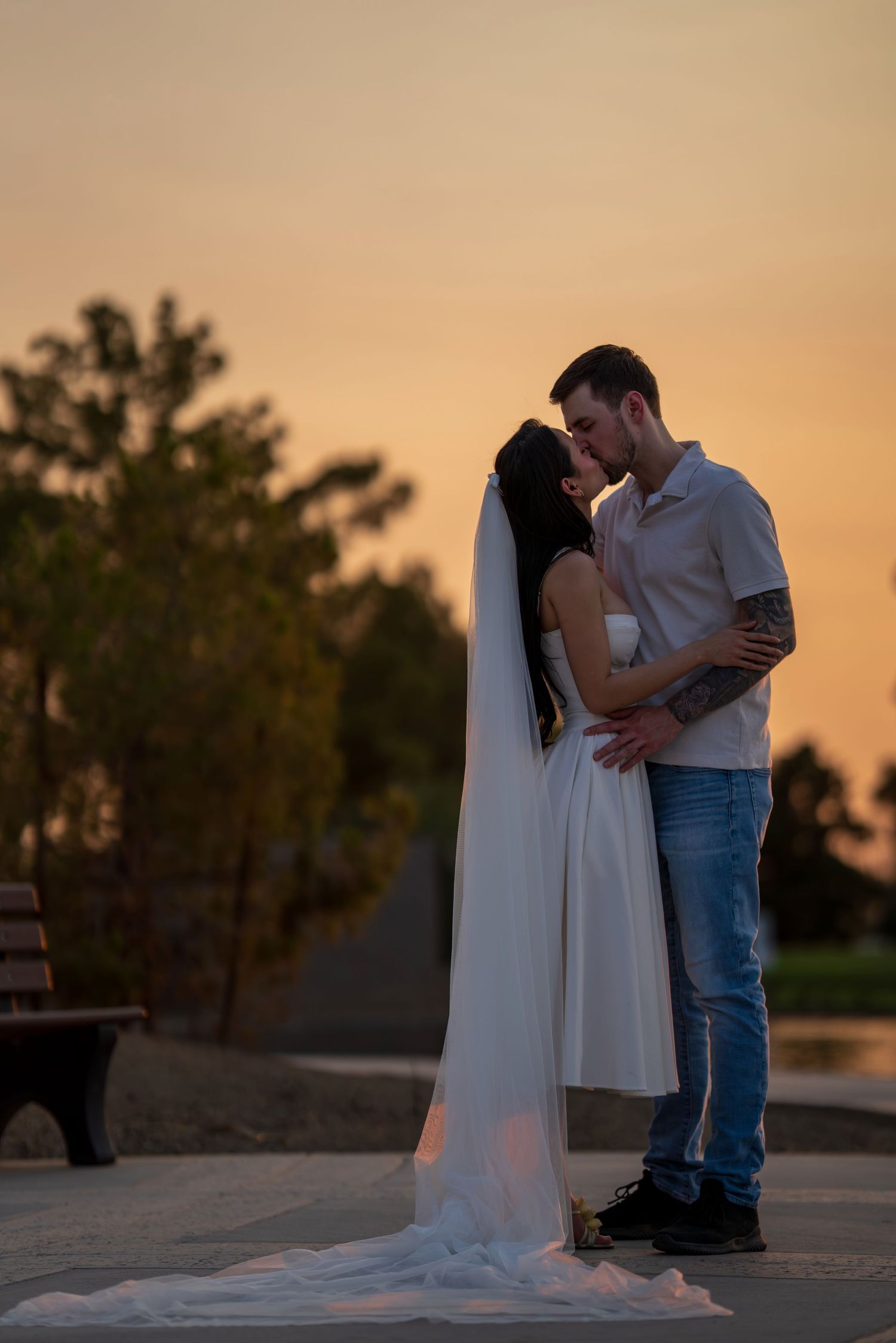 A bride in a white gown and veil kisses her groom at a stunning sunset.