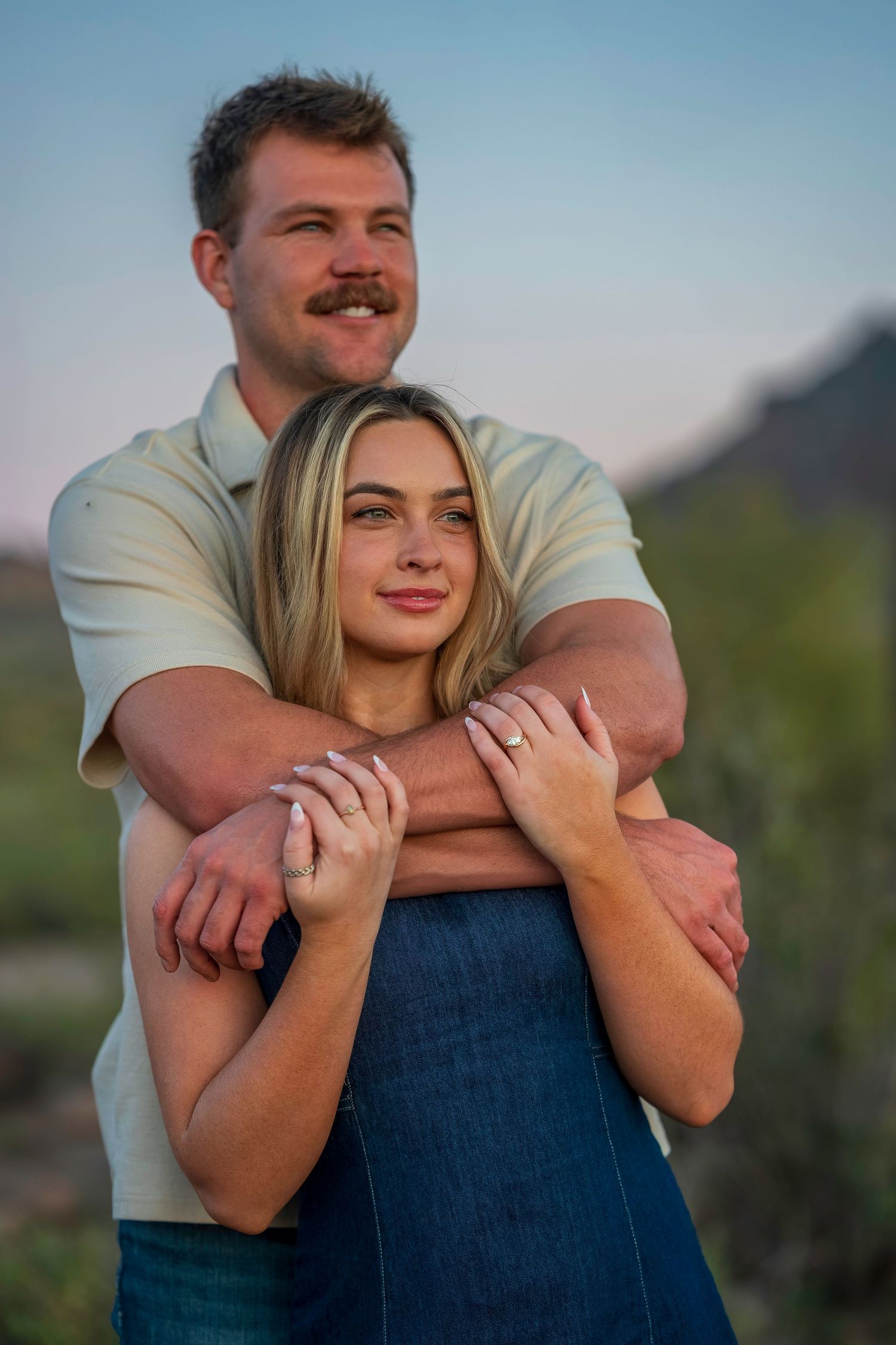 Couple embracing at sunset in Arizona desert, man wrapping arms around blonde woman showing engagement ring.
