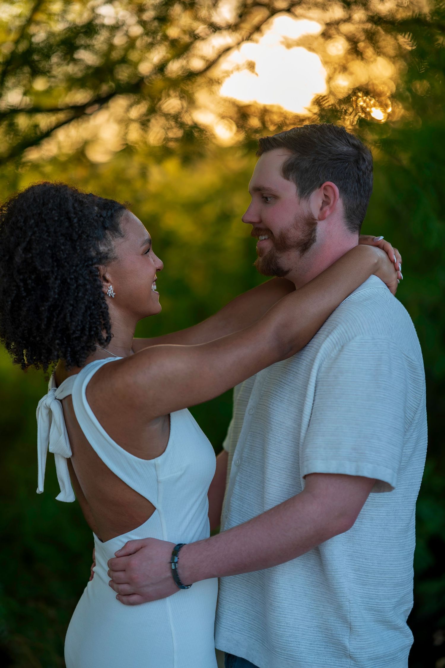 Happy interracial couple gazing at each other smiling, embracing outdoors during golden sunset.