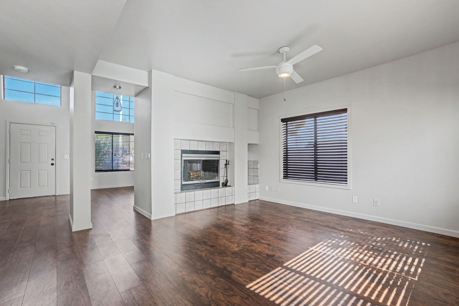 Bright living room with hardwood floors, white tile fireplace, ceiling fan, and large windows letting in natural light.