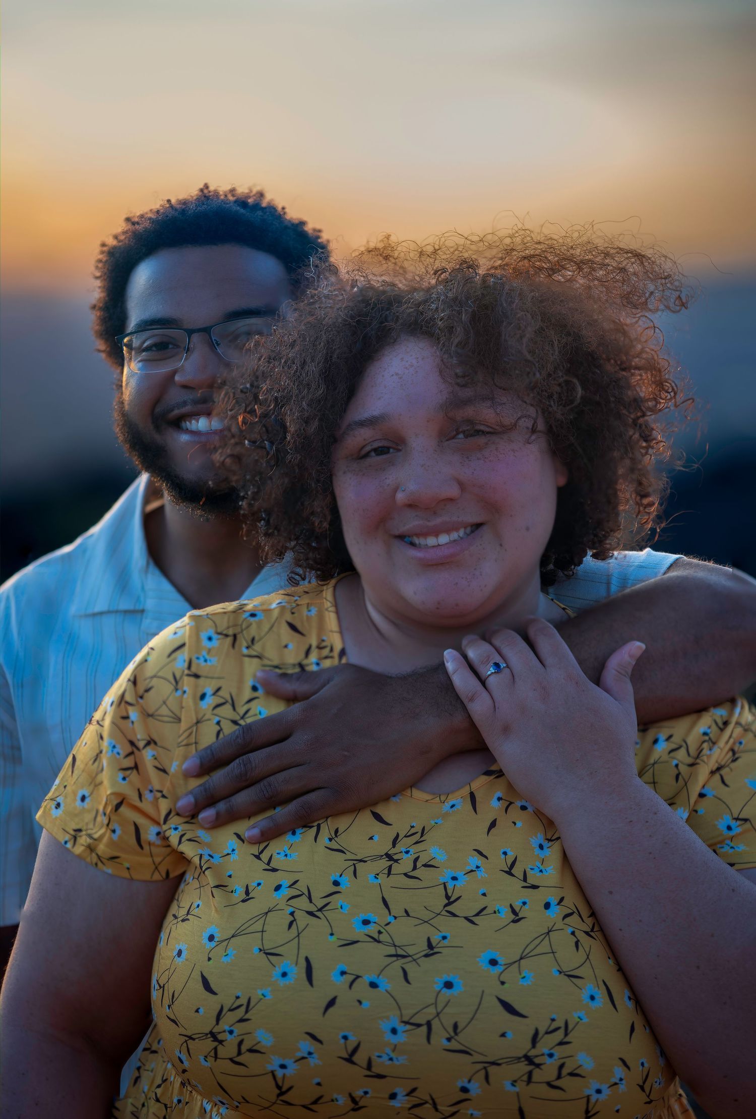 Smiling couple at sunset, woman in yellow floral dress with curly hair, man in white shirt with glasses embracing her.