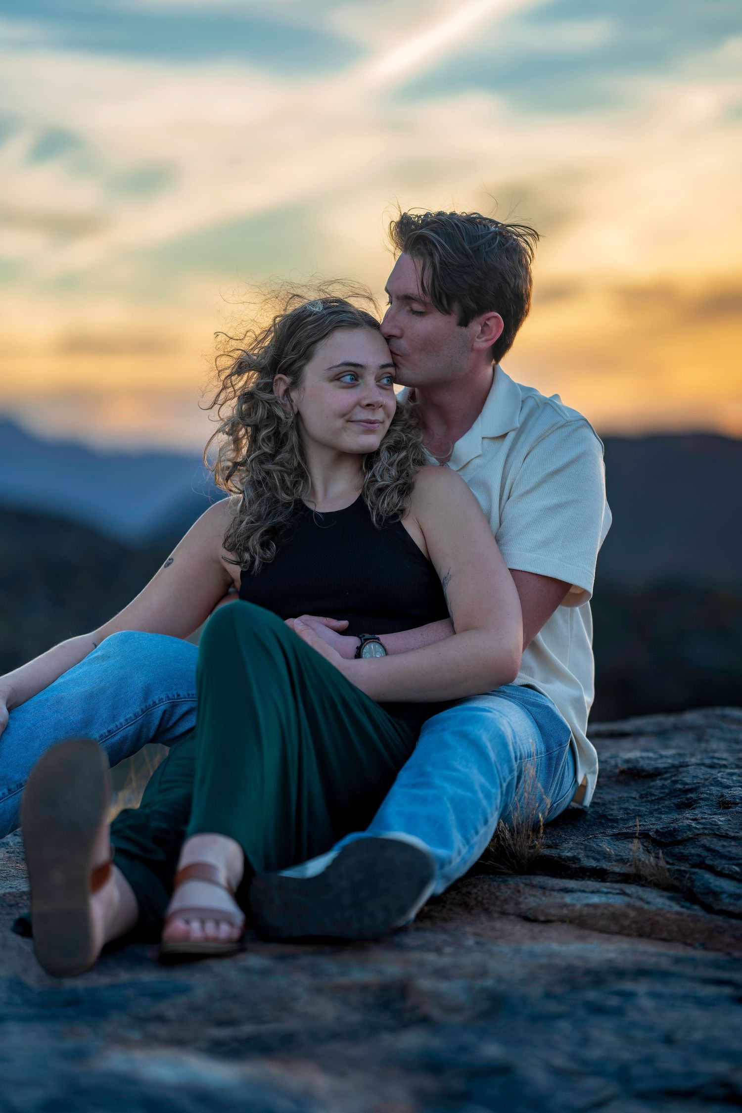 Couple sitting together on a rock at sunset, man kissing womans temple.