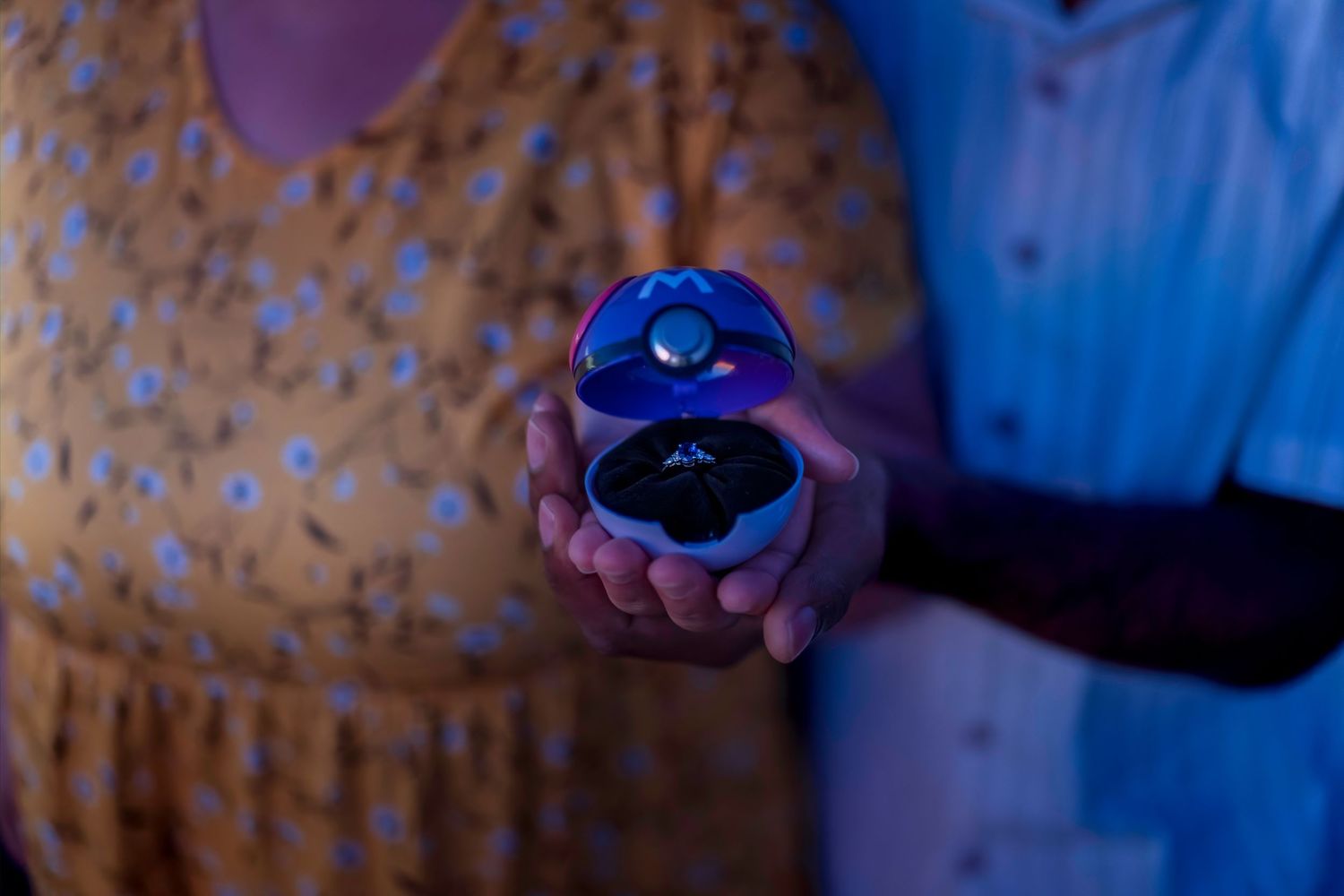A couple holds open a blue velvet ring box with a diamond engagement ring inside, lit with a glowing blue light.