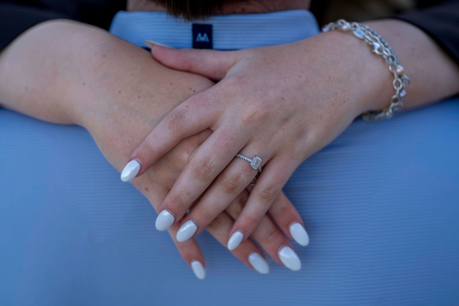 Close-up of woman's hands with white oval nails, diamond engagement ring, and silver chain bracelet resting on blue fabric.