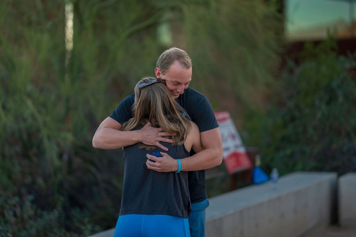 A man and woman share a warm embrace outdoors, surrounded by lush green trees in soft evening light.