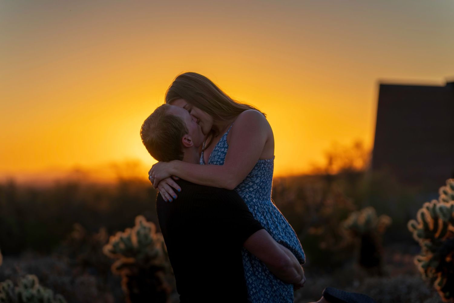 Couple sharing passionate kiss silhouetted against vibrant orange Arizona desert sunset with cacti.