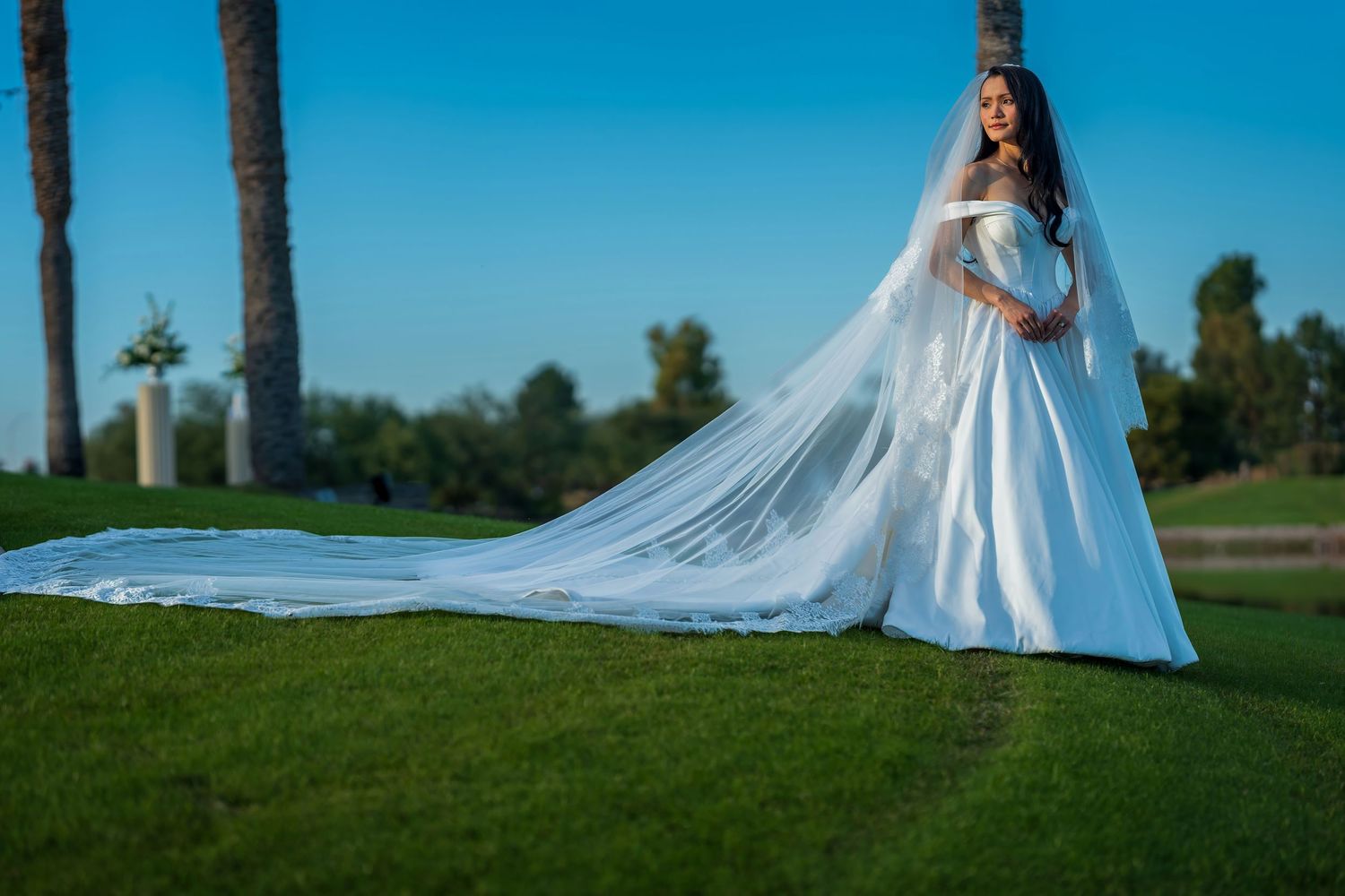 A bride in a white gown walks gracefully across a green field with her veil flowing in the breeze.