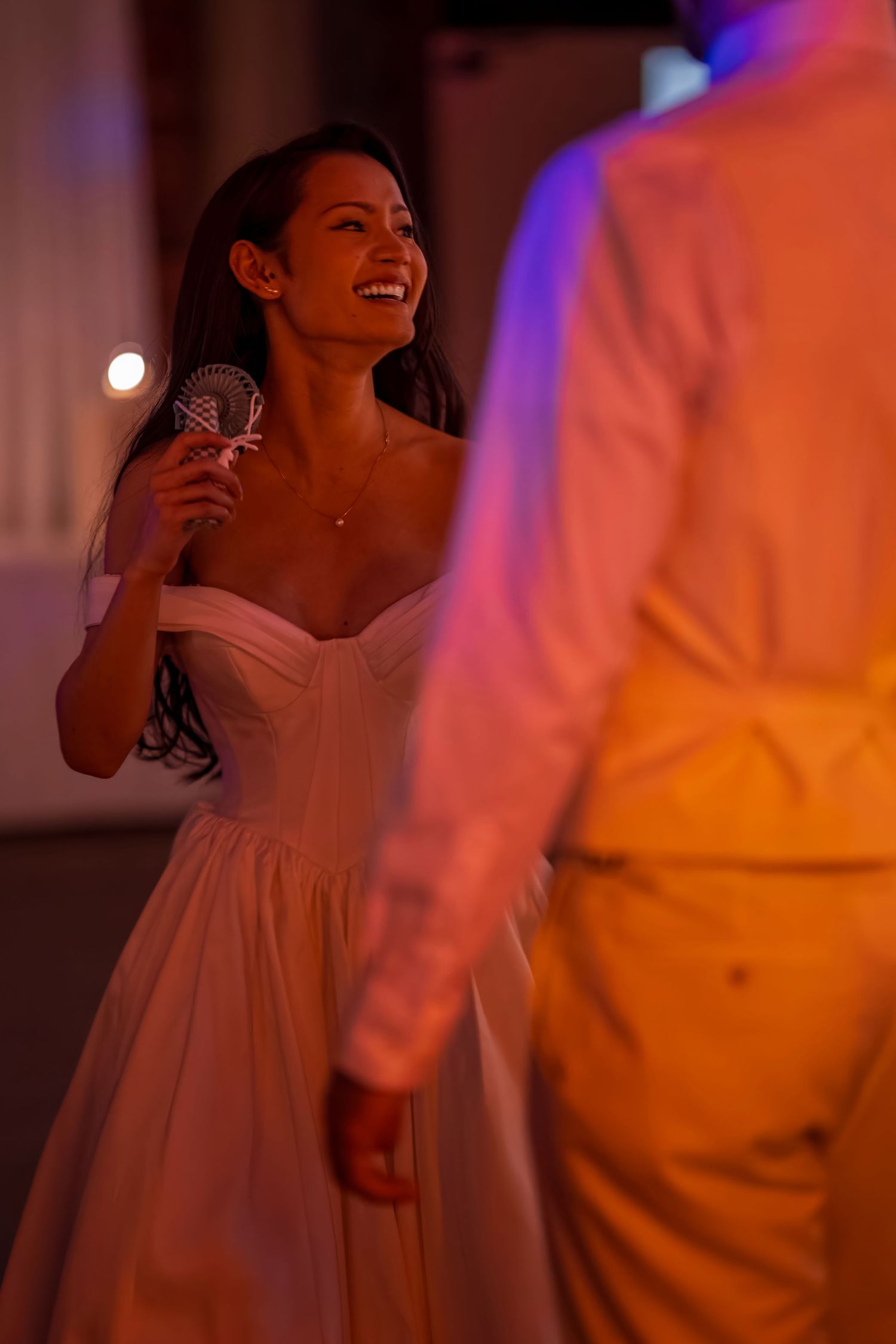 A bride in a white dress laughs while holding a microphone at a reception.