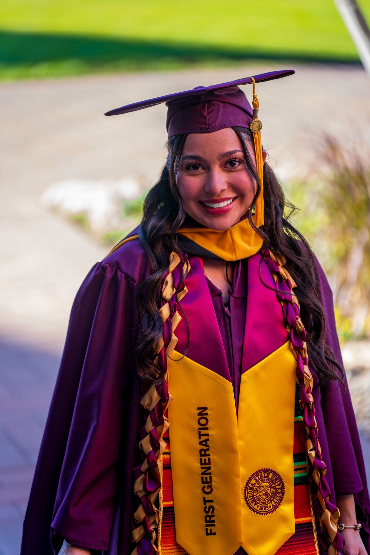 Graduate in maroon academic regalia with yellow stole and braided cord poses outdoors.