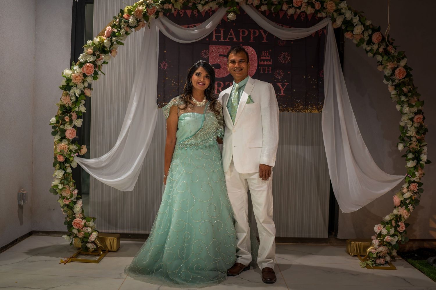 A couple poses under an elegant floral arch with white draping, wearing mint green and white formal attire.