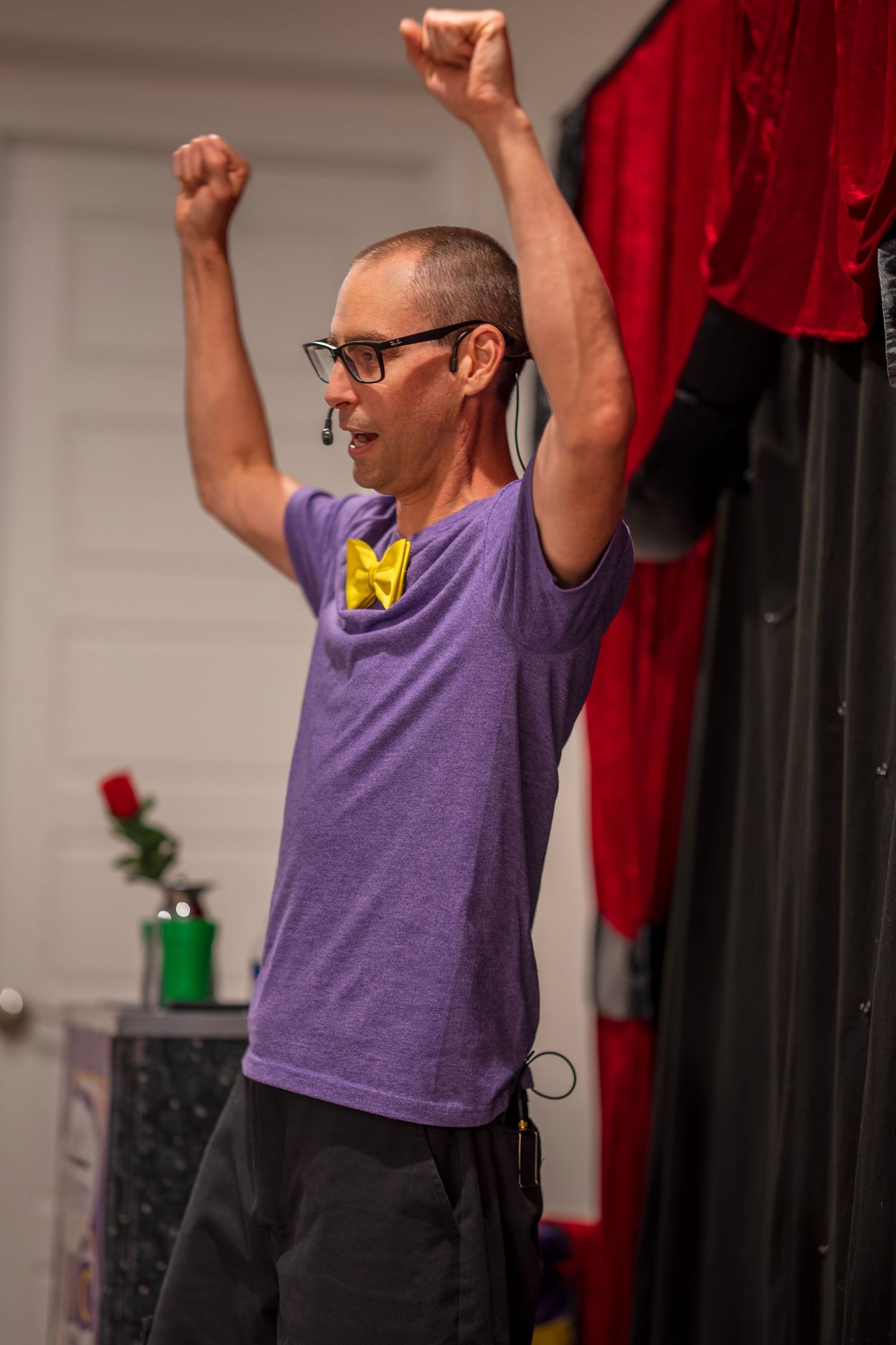 Performer in purple t-shirt and yellow bowtie raises arms enthusiastically during show.