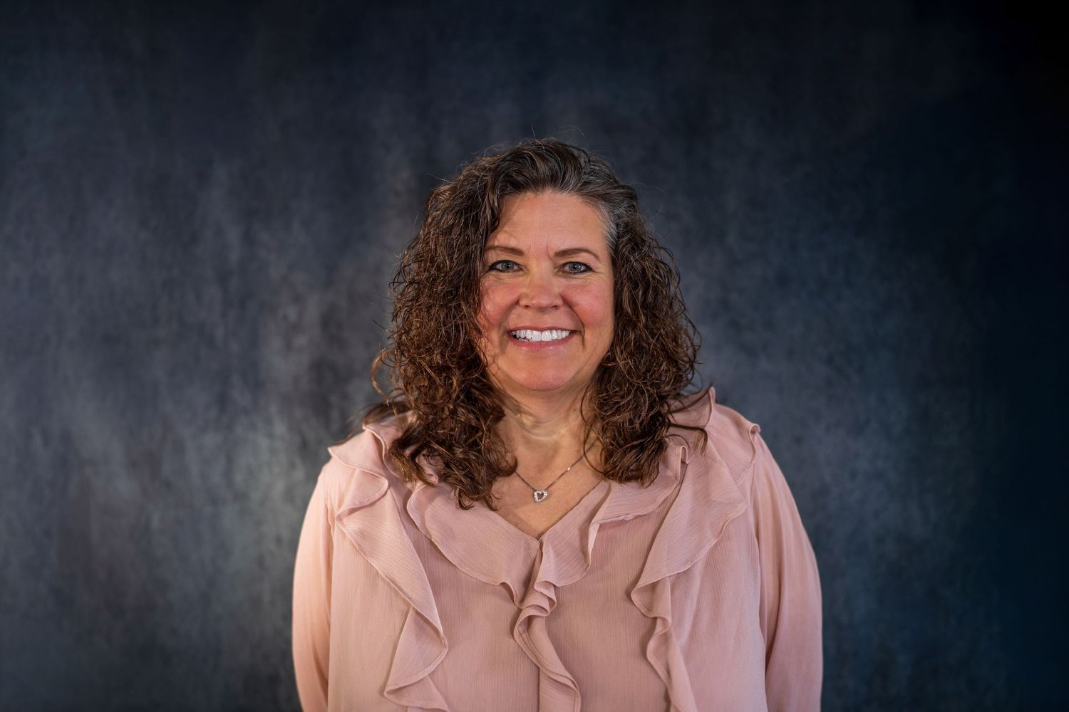 Professional portrait in pink blouse showing joyful expression against dark background.