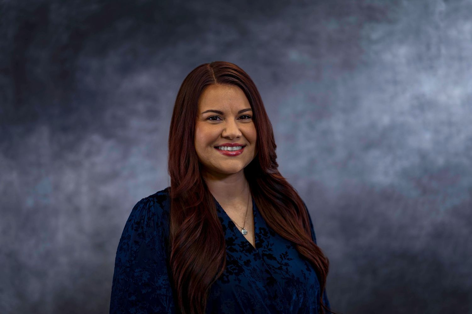 Professional headshot of employee with long auburn hair wearing navy blue top against dark background.