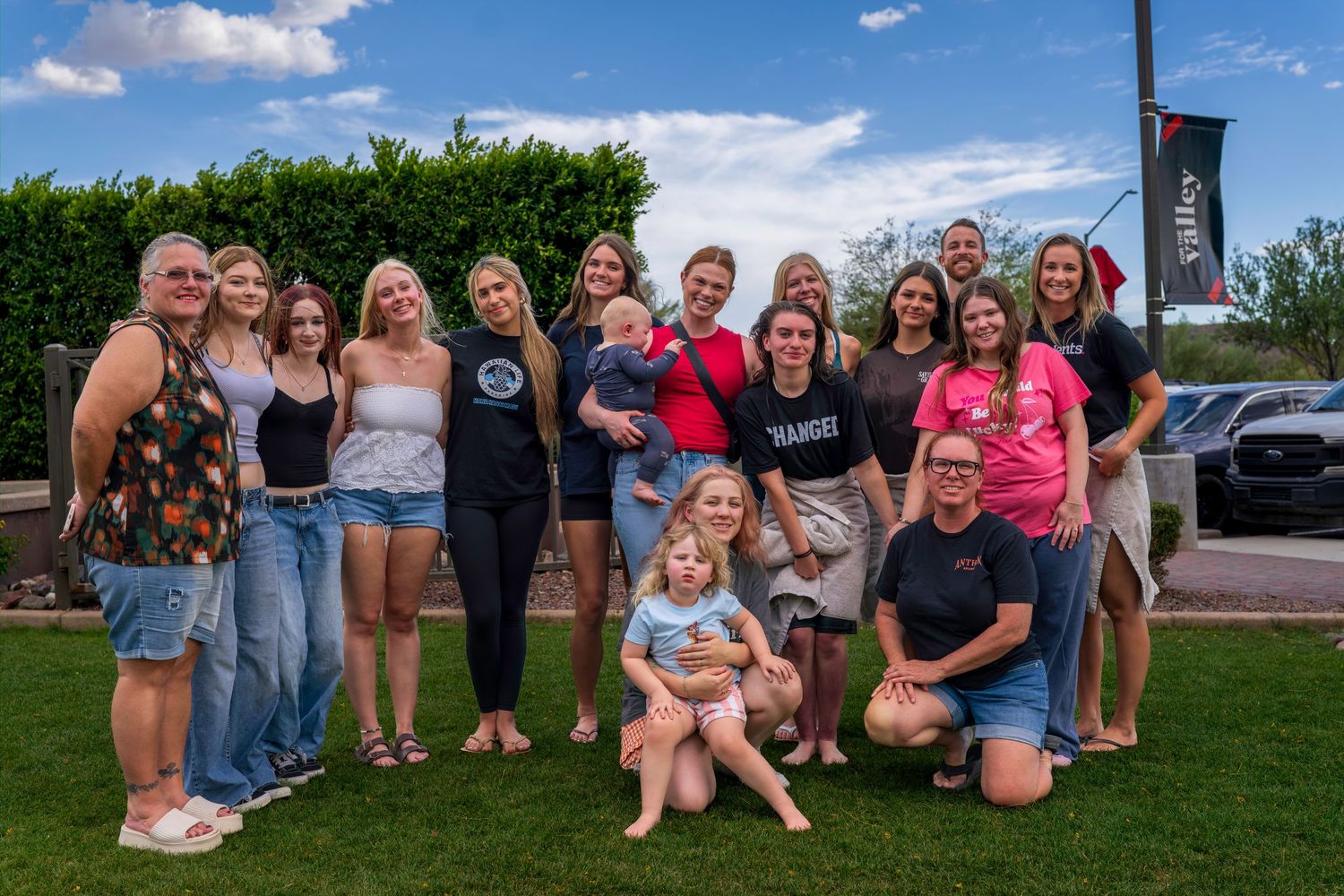 Large group of smiling people posing together outdoors on a sunny day on a green lawn.