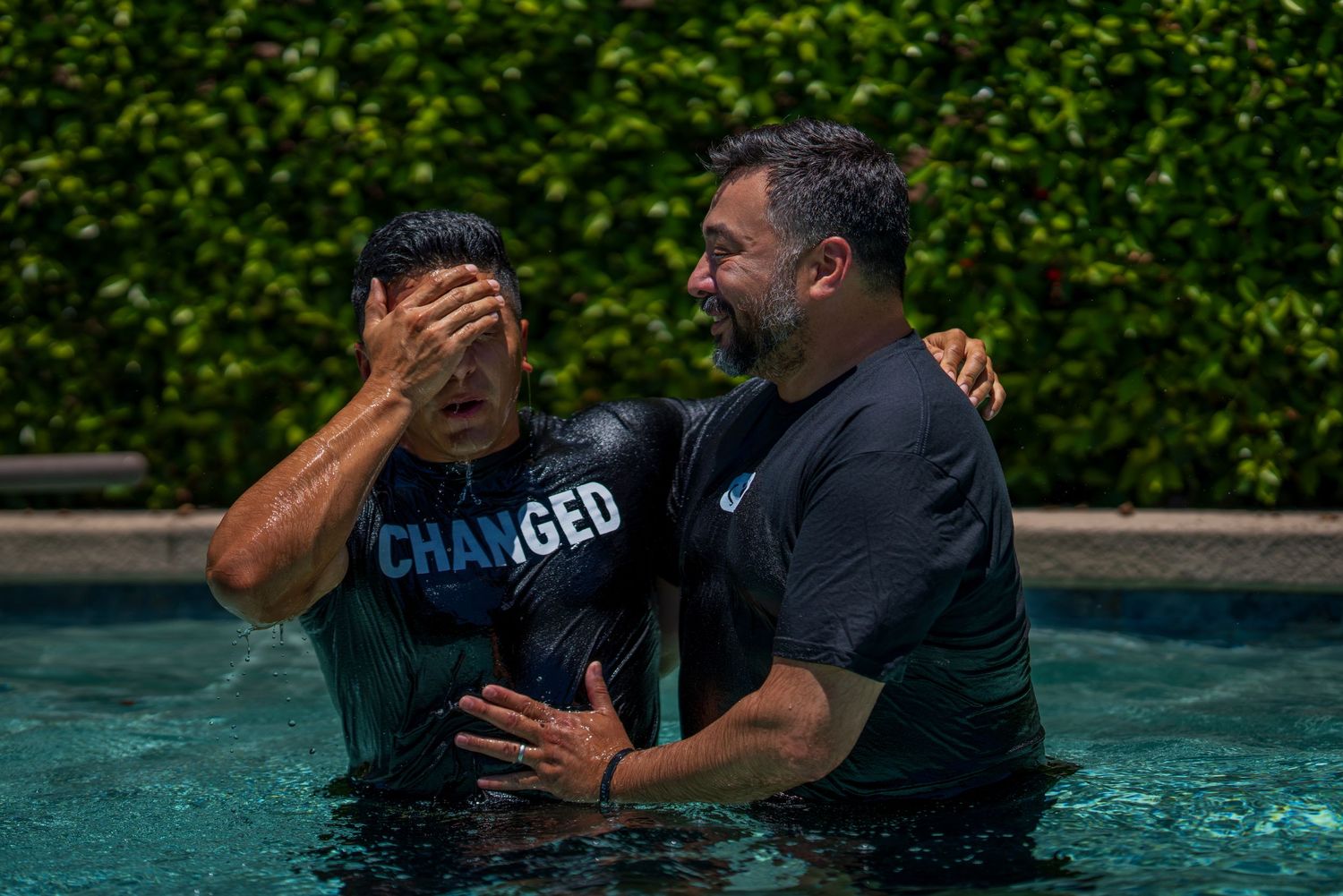 Man covering his face during a baptism in a pool, supported by another man wearing a dark shirt.