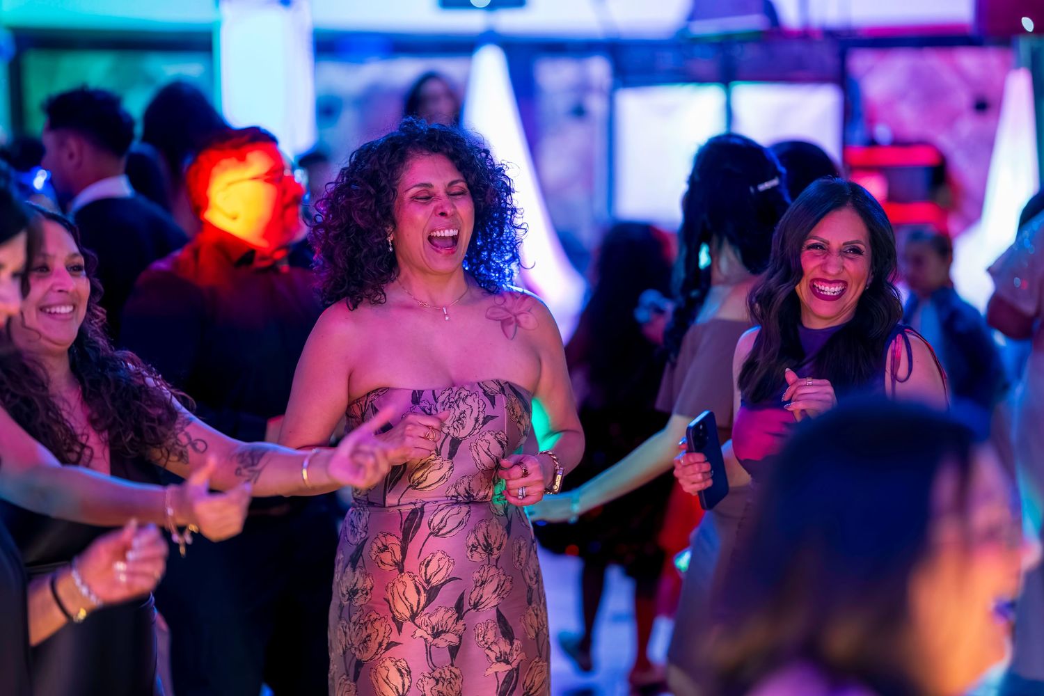 Women laughing and dancing together at a lively party with colorful lights on the dance floor.