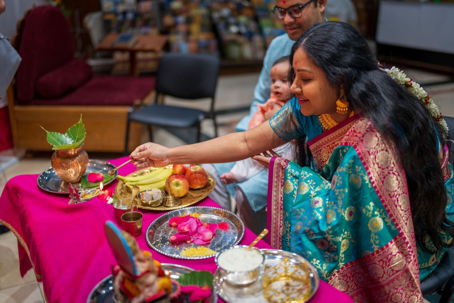 Woman in teal and red saree performing Hindu puja ritual with offerings on pink cloth-covered table.