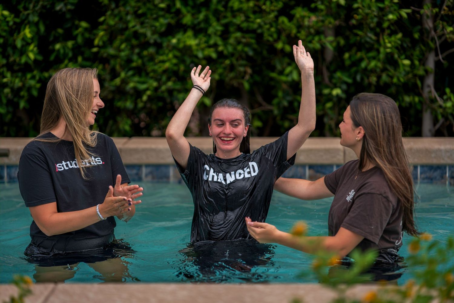 Three young women celebrating a baptism in a pool, with one raising her arms joyfully wearing a ‘Changed’ shirt.