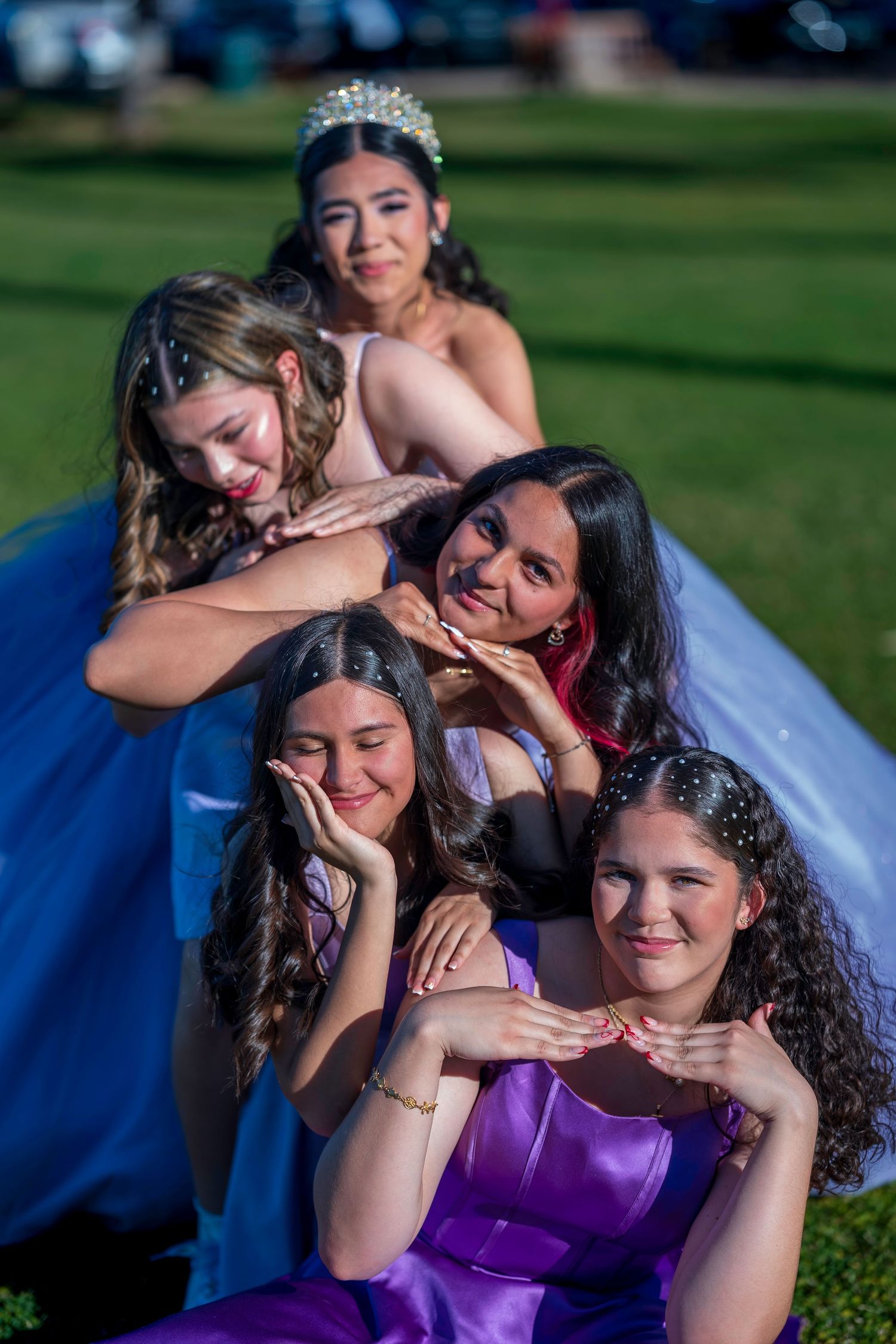 Five young women in colorful formal gowns pose together outdoors on grass during a quinceañera celebration.