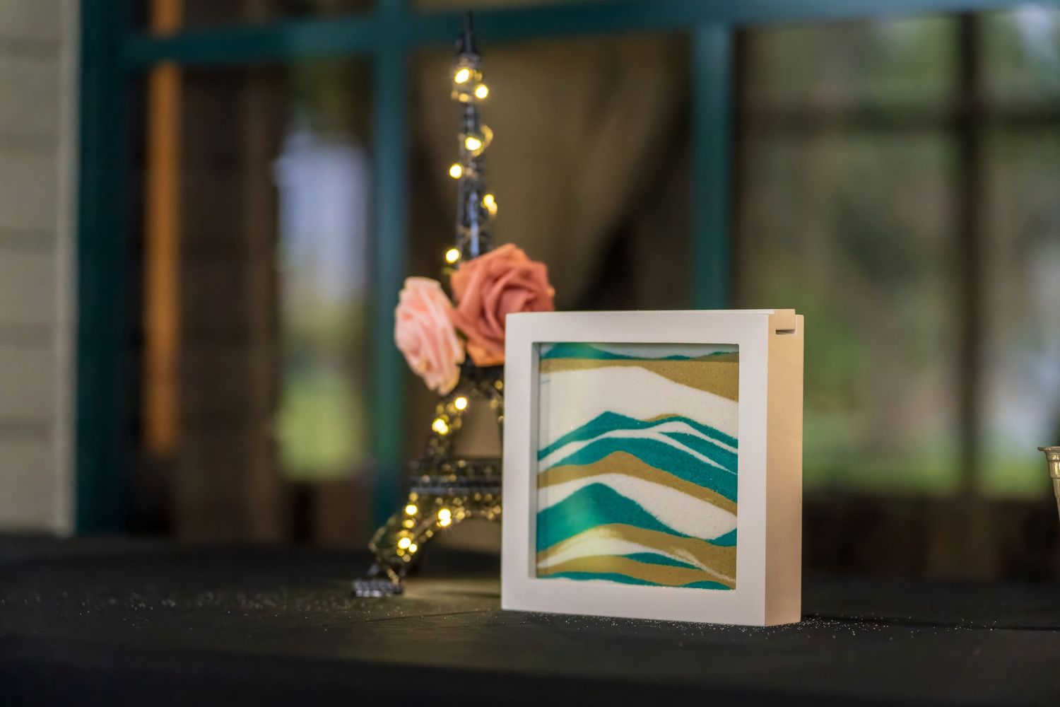A framed layered paper art piece with teal and gold mountain waves displayed on a table with fairy lights and pink roses.