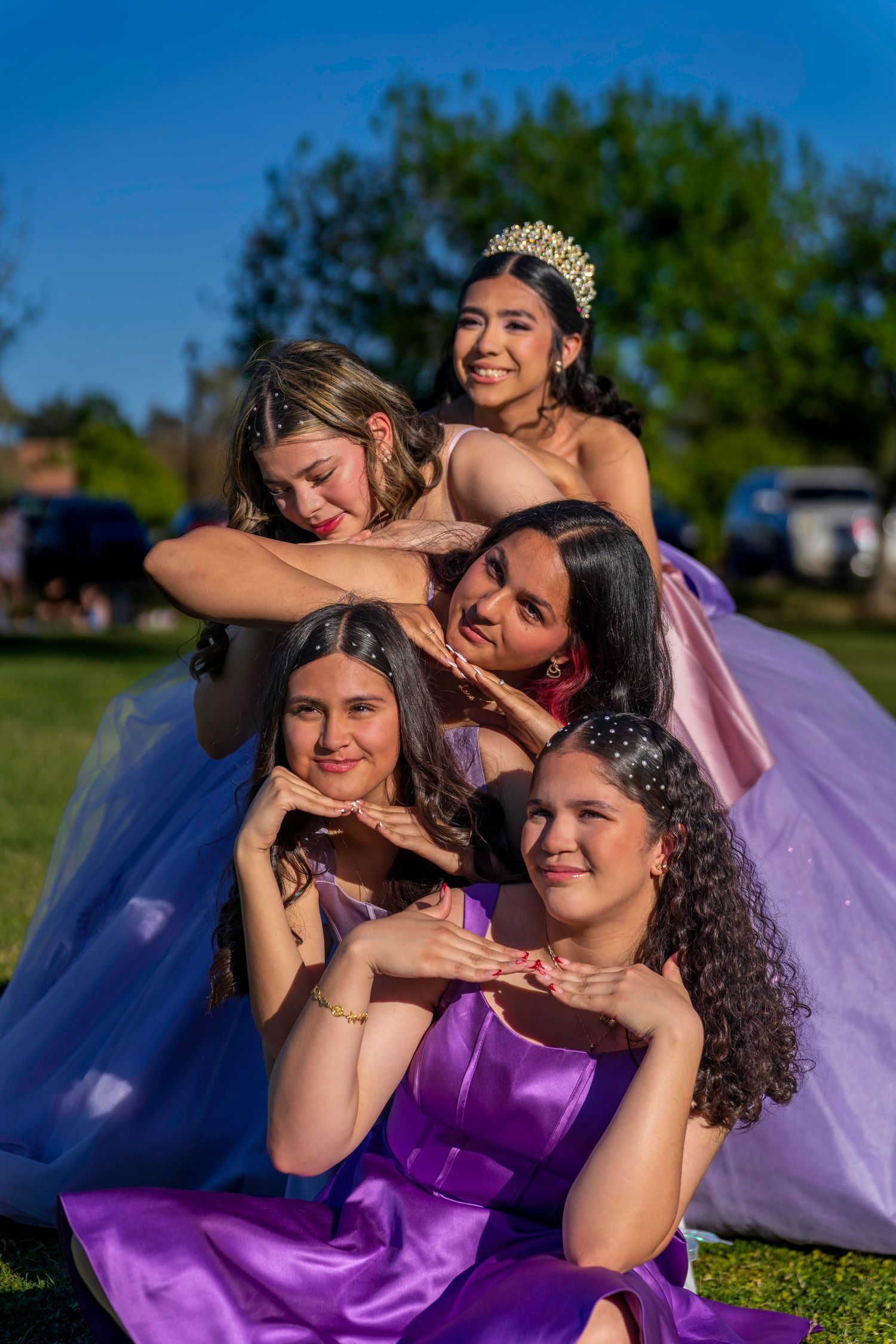 Five young women in purple and pink formal gowns pose together outdoors during a quinceañera celebration.