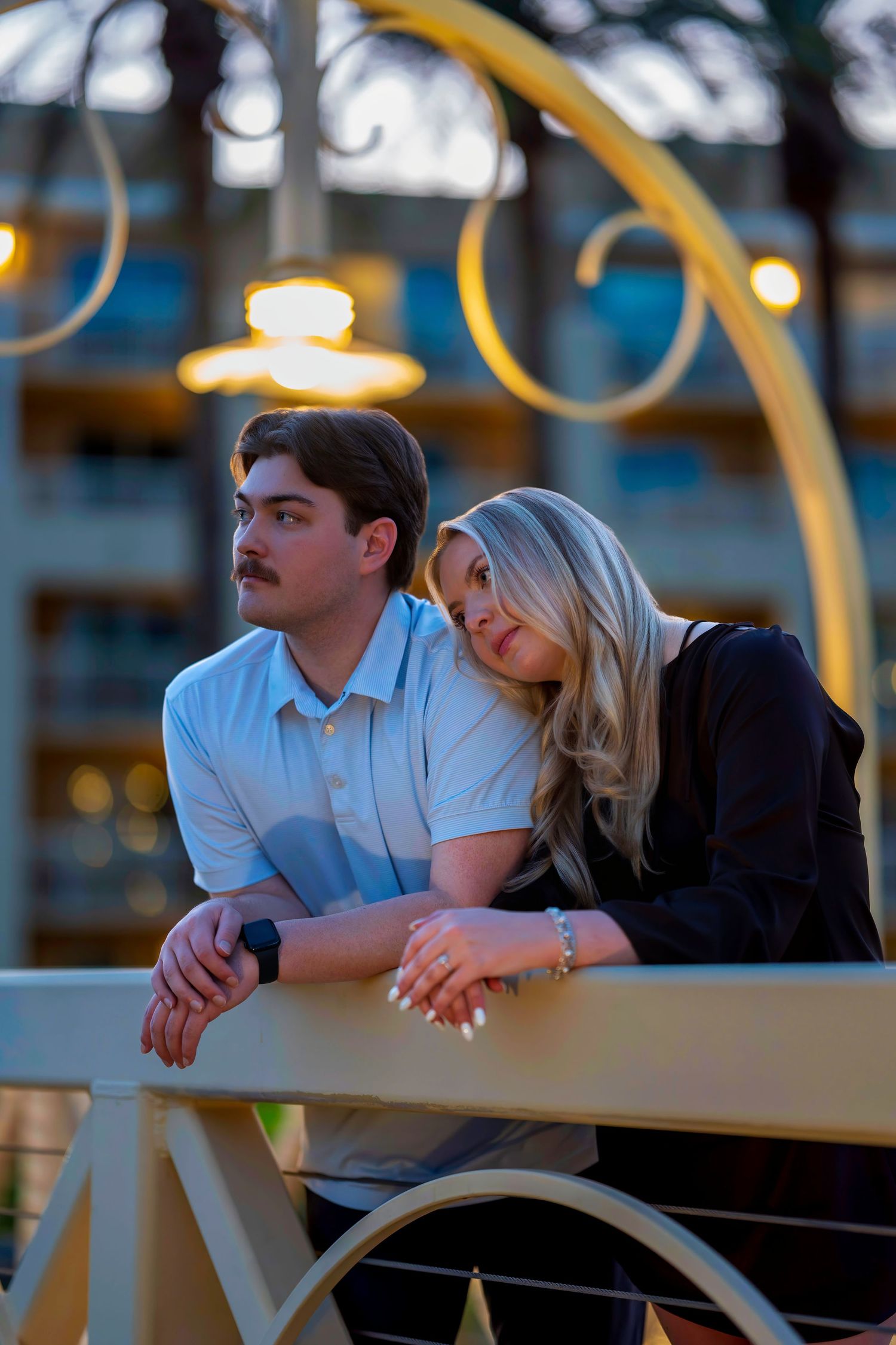 Engaged couple leaning on a railing under decorative lights at a resort during evening hours.