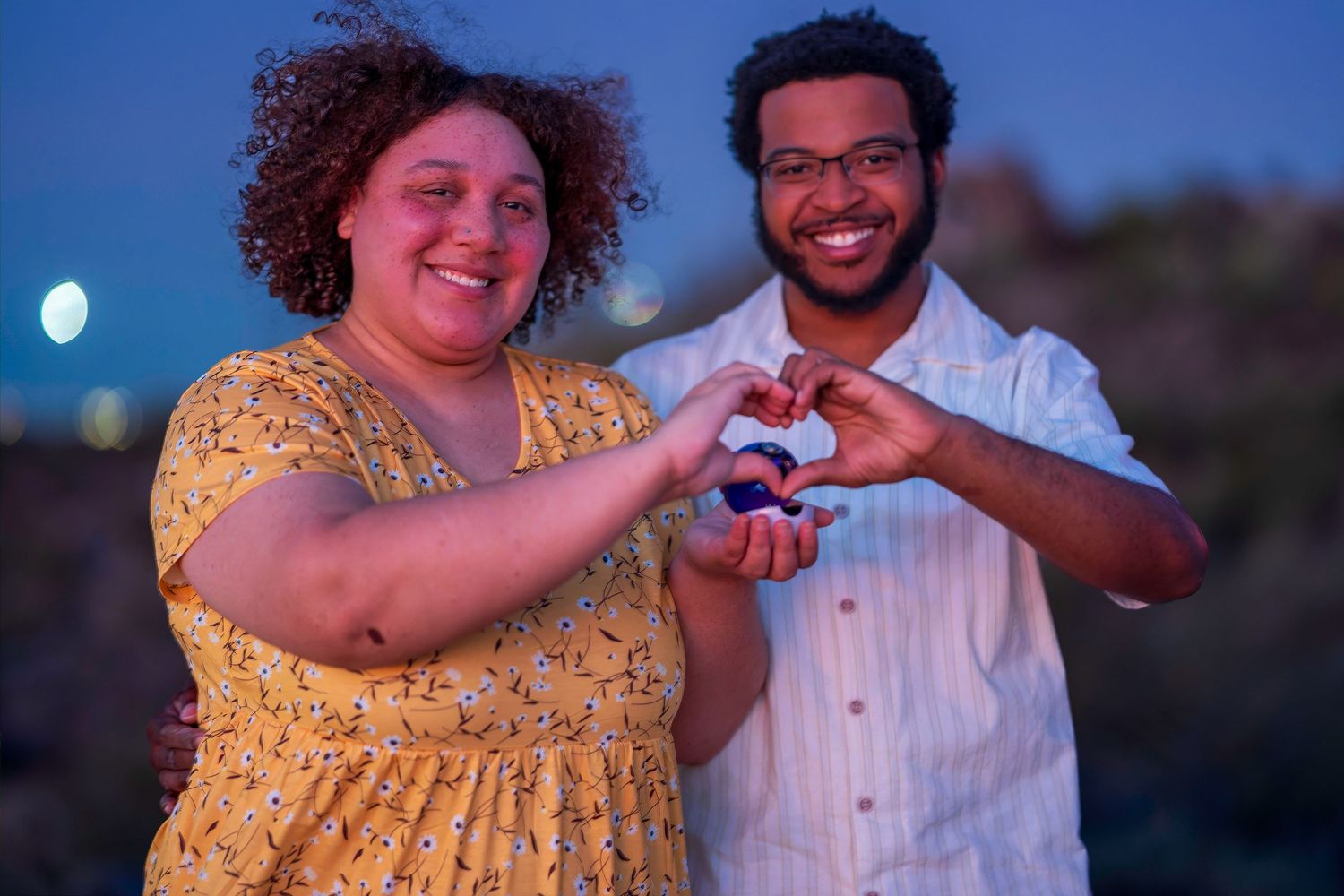 Happy couple forms a heart shape with their hands together, smiling at camera during twilight outdoors.