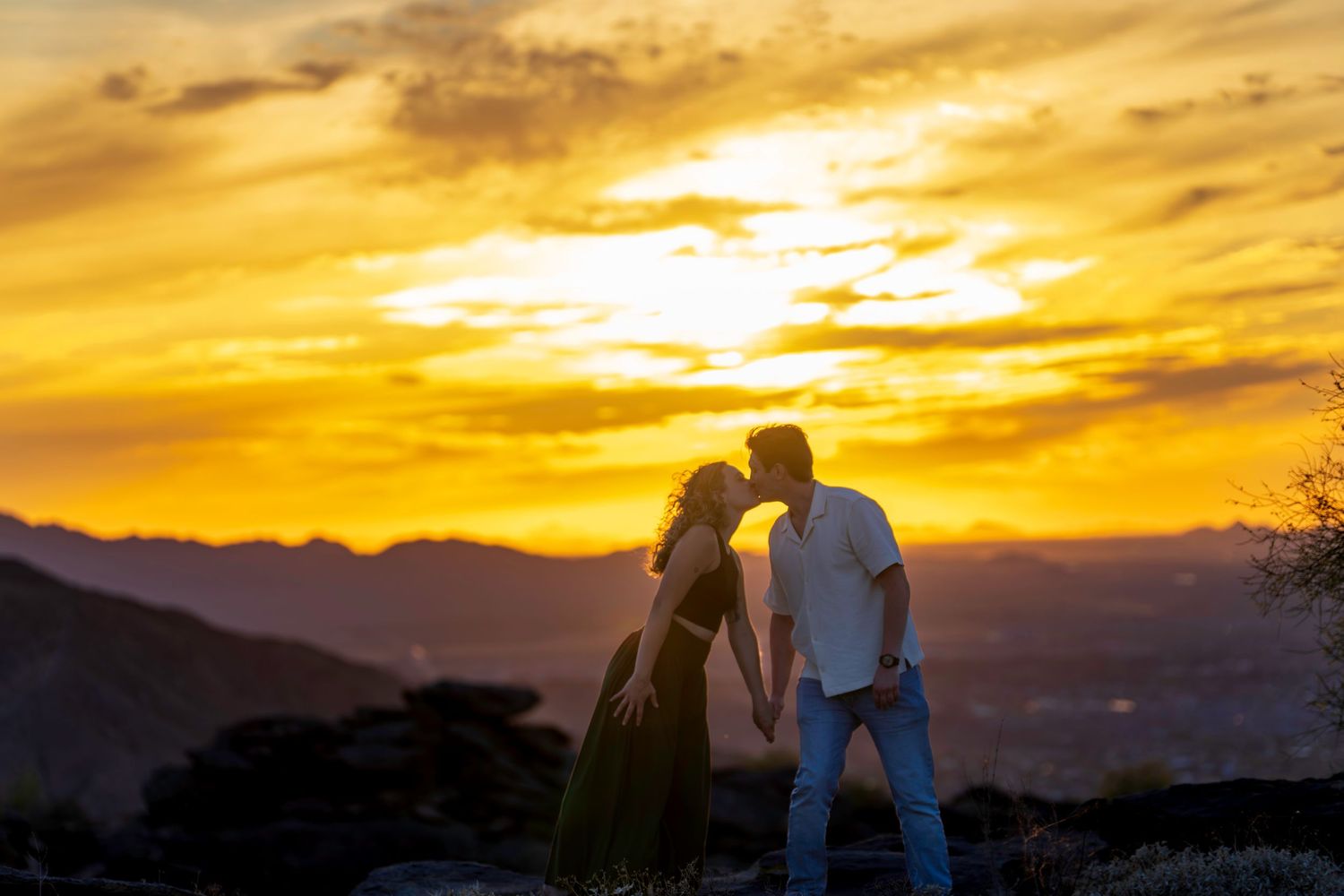 Couple kissing in silhouette against a vibrant golden sunset over mountains.