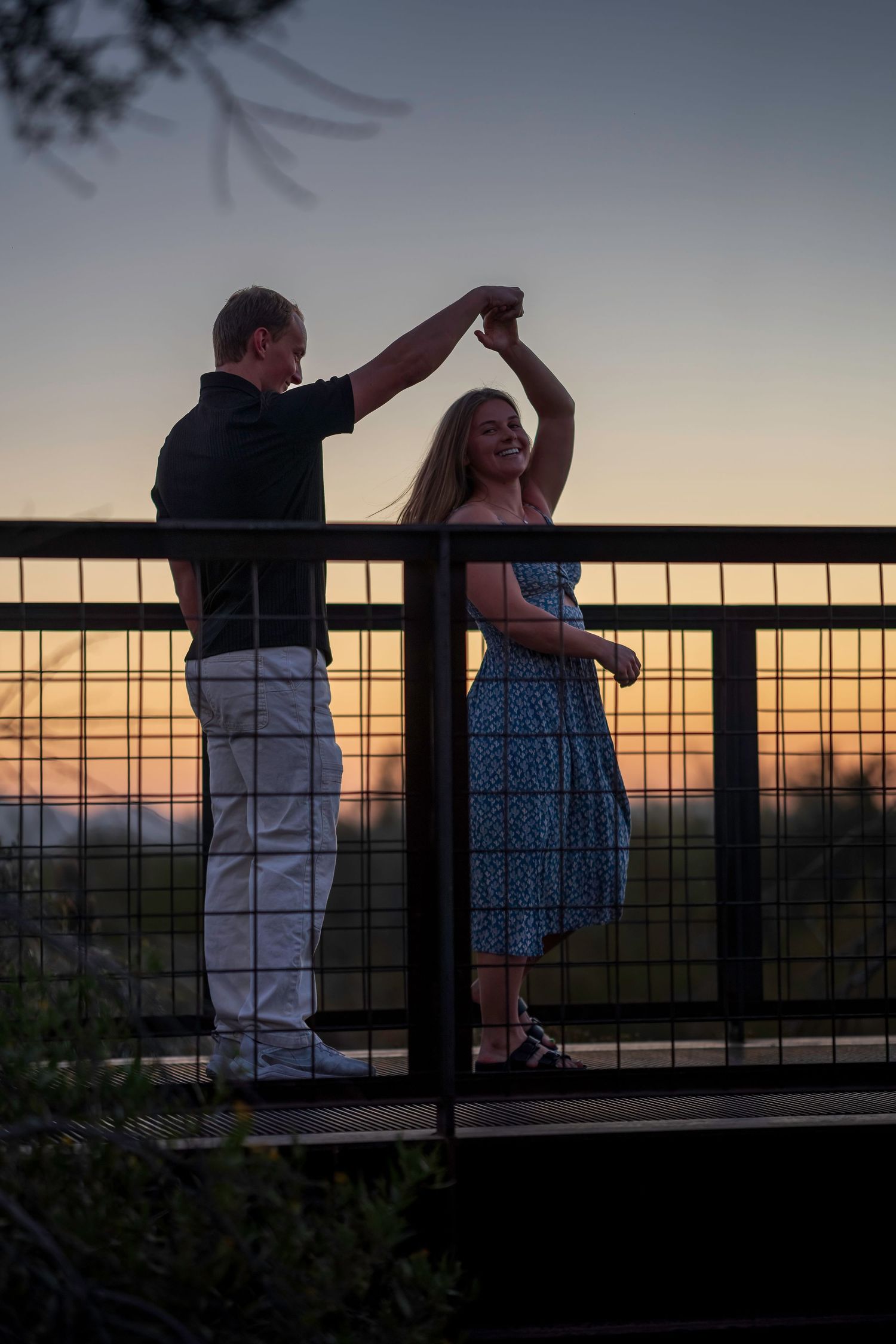 Couple dancing at sunset on overlook bridge, man spinning woman in blue dress against golden horizon.