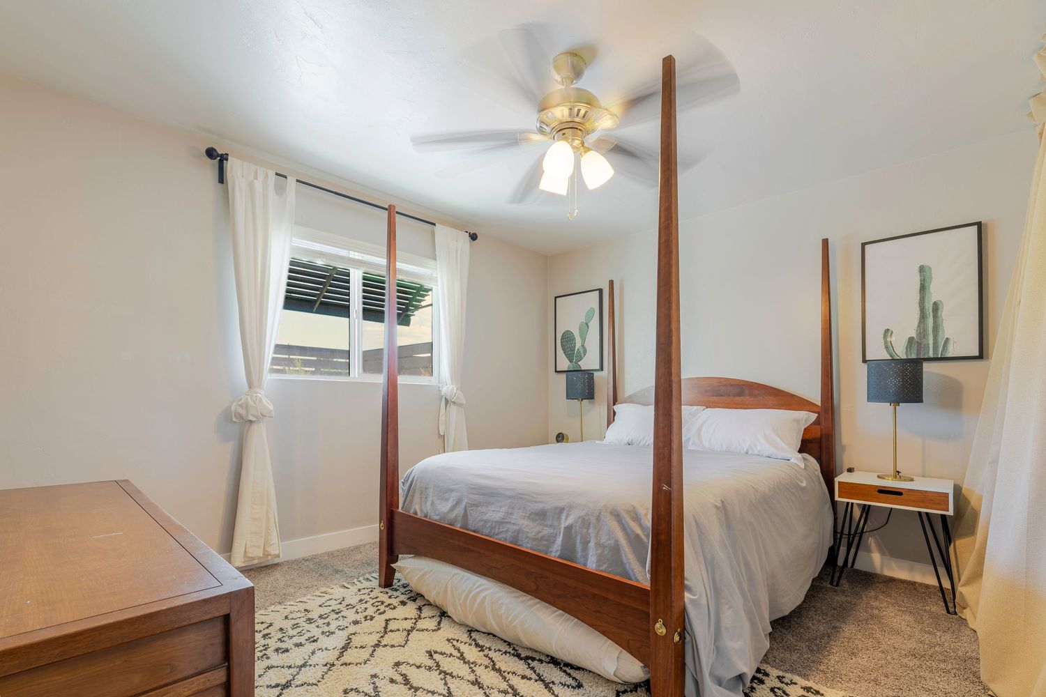 Cozy bedroom with four-poster bed, ceiling fan, patterned rug, and cactus decor beside a sunny window.