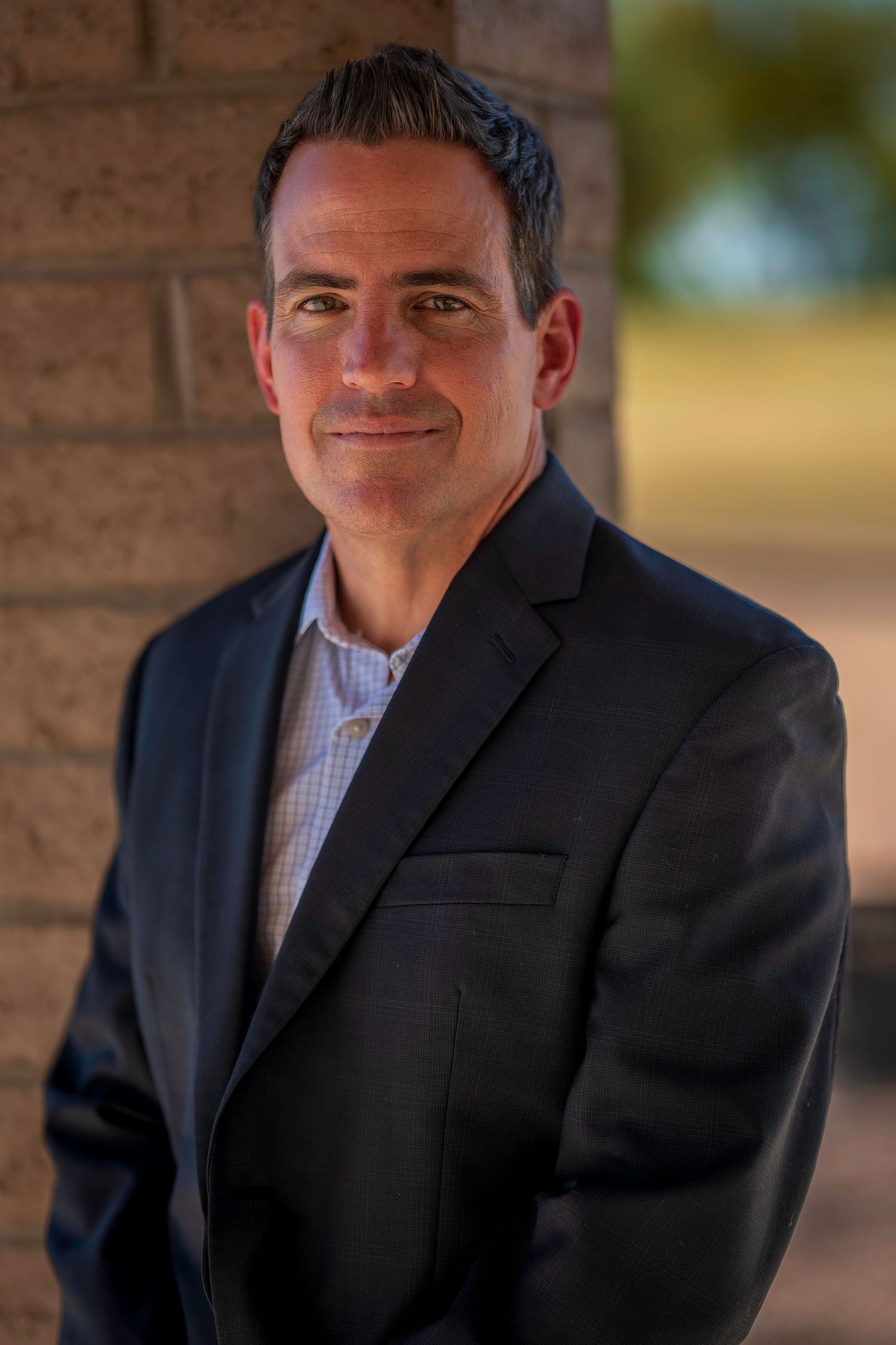 Professional man in dark blazer smiling confidently outdoors near brick wall in warm natural light.