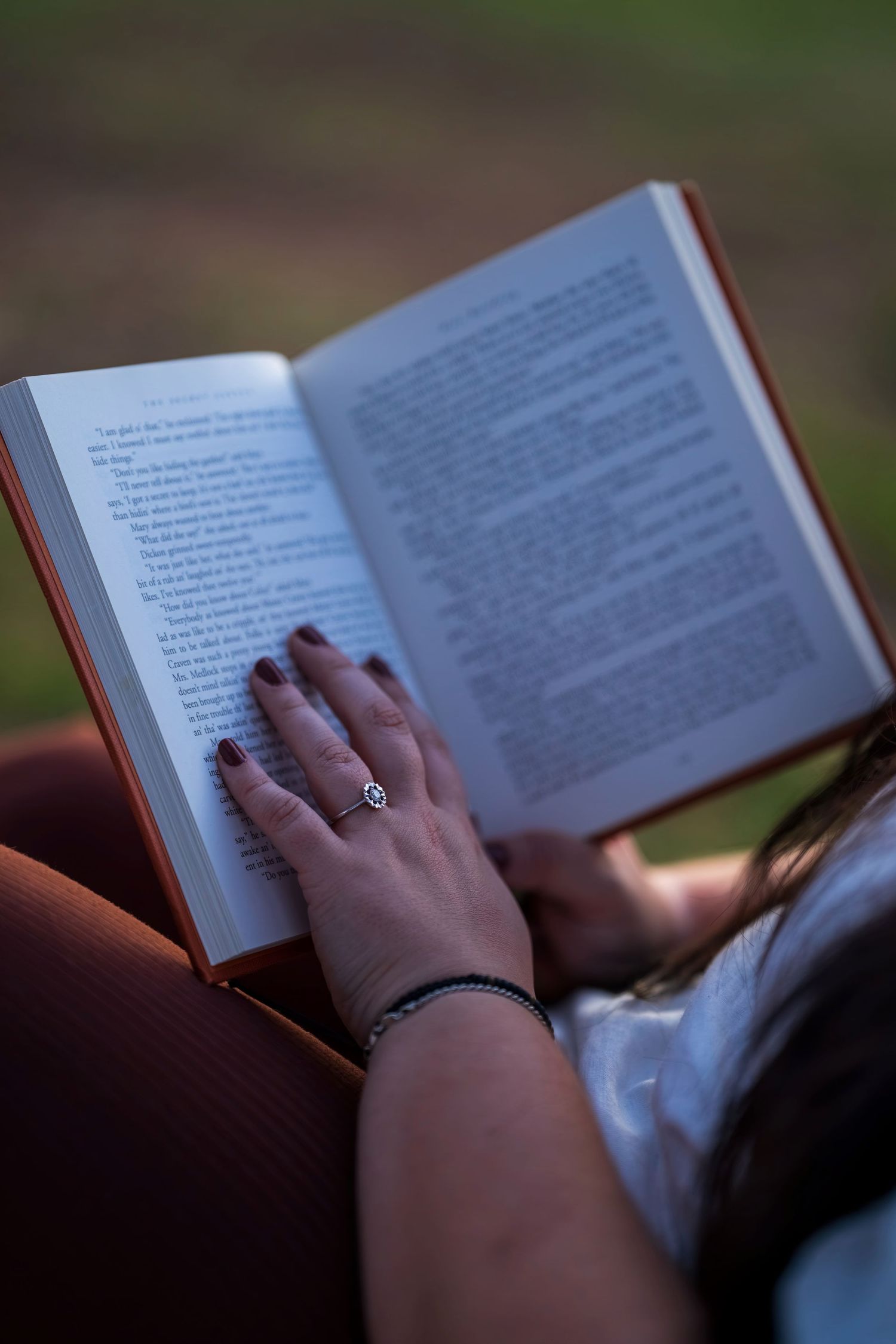 A woman reading an open book outdoors, wearing a decorative ring and bracelet on her hands.