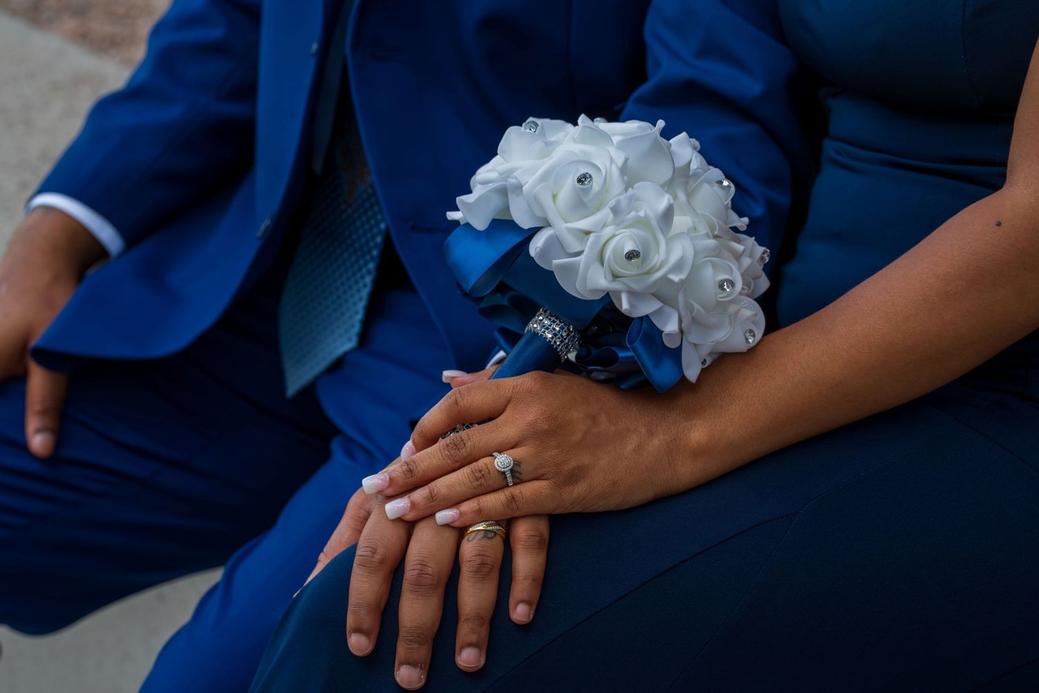 Close-up of couple in blue wedding attire holding hands with white rose bouquet and diamond engagement ring.