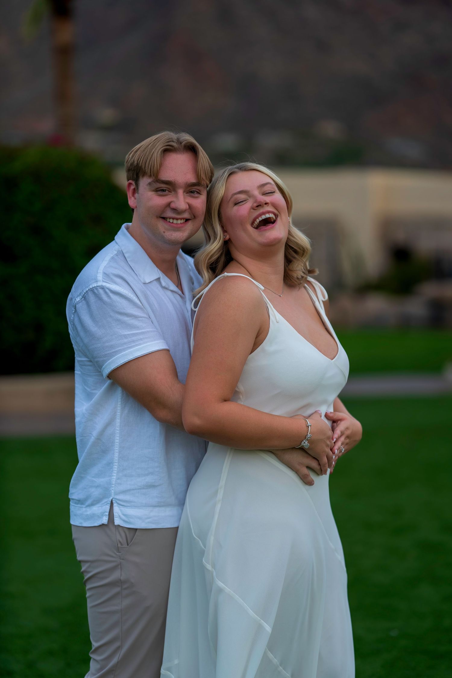 A laughing couple in white outfits embrace on a green lawn with desert mountains in the background.