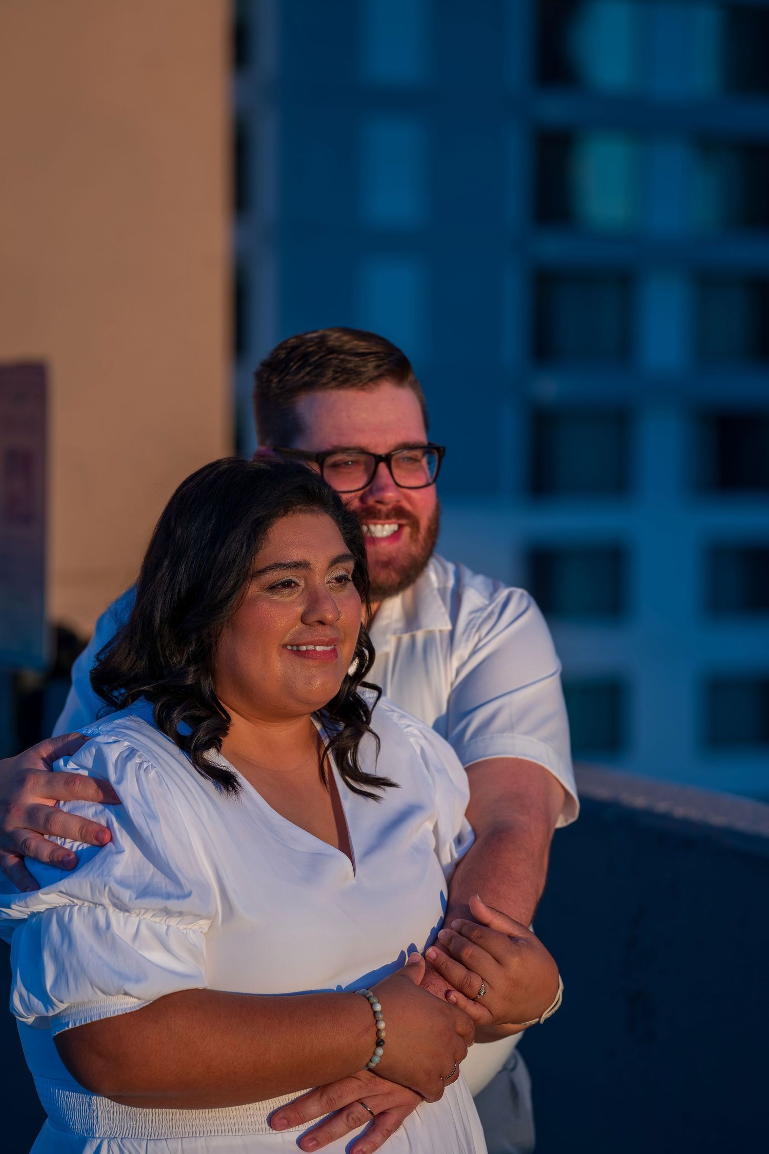 Couple in white outfits embracing on a rooftop at night, smiling warmly with city buildings in the background.