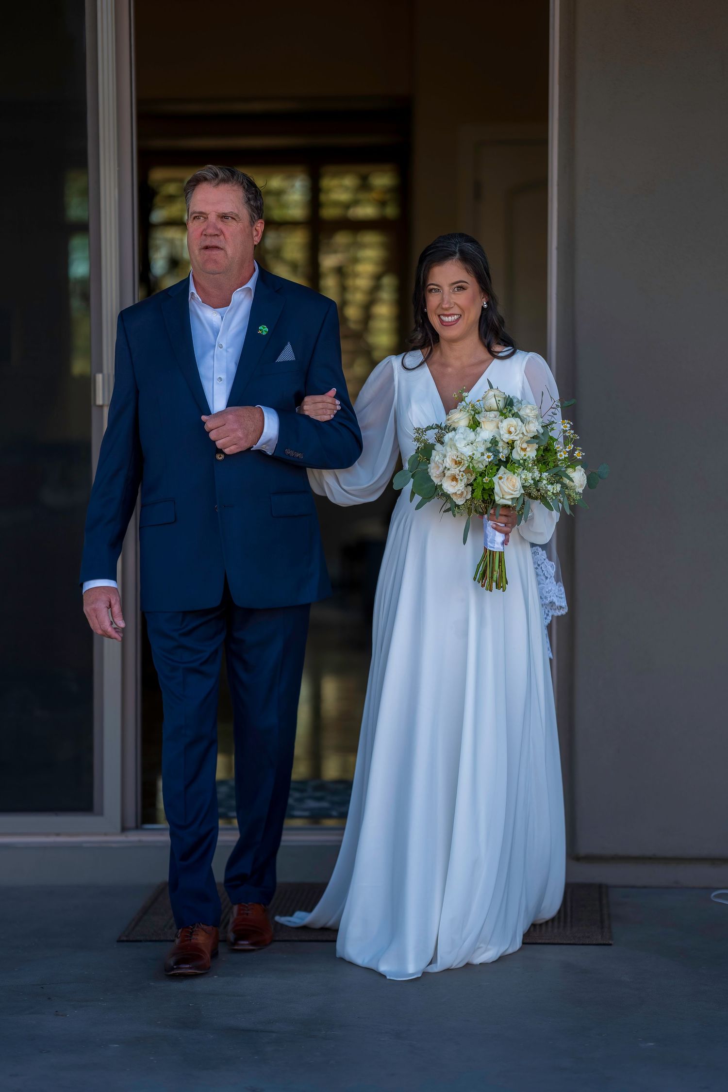 Bride in white gown holding bouquet walks arm-in-arm with man in navy suit at wedding ceremony.