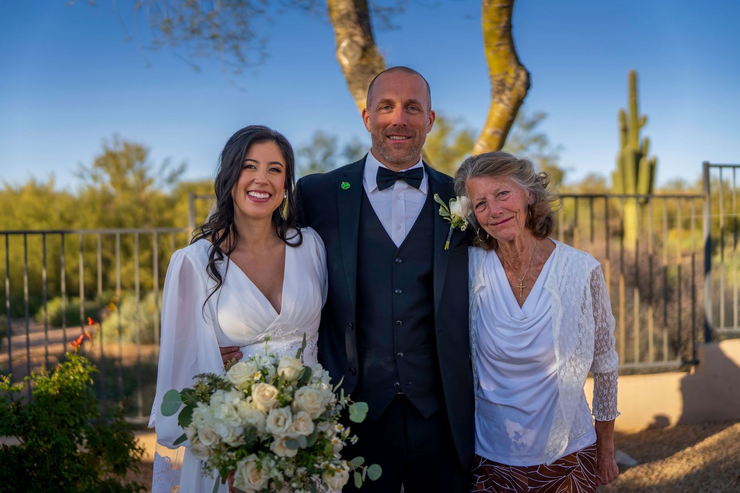 Bride and groom pose with older woman guest at outdoor desert wedding with saguaro cactus backdrop.
