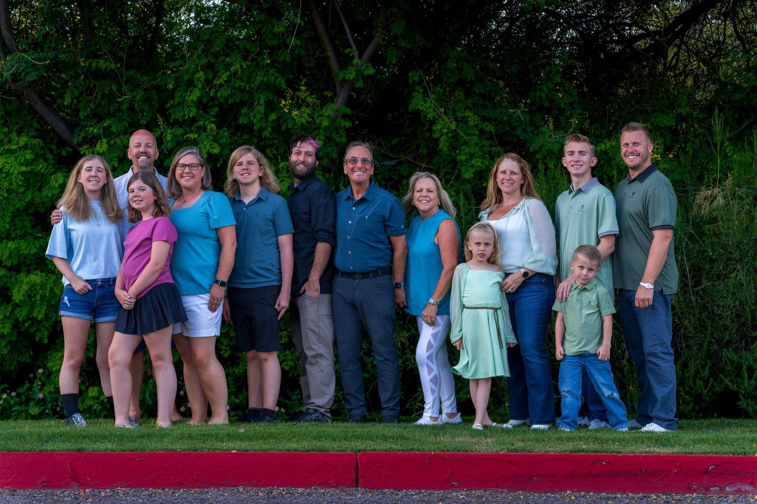 Large extended family group portrait of about 13 people posing together outdoors near green trees.