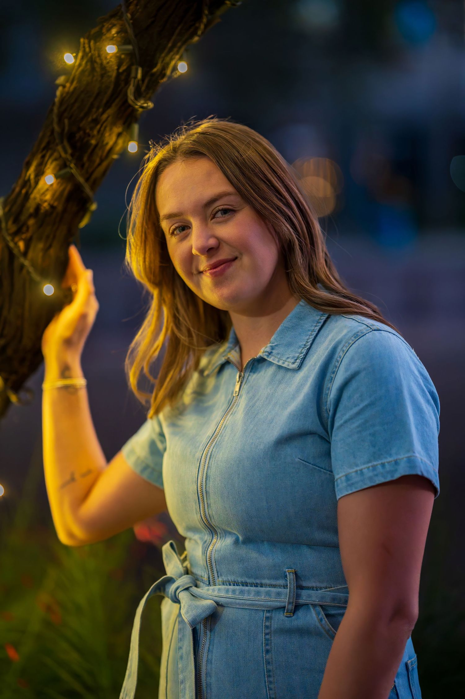Young woman in denim dress smiling outdoors at night with warm string lights glowing behind her.