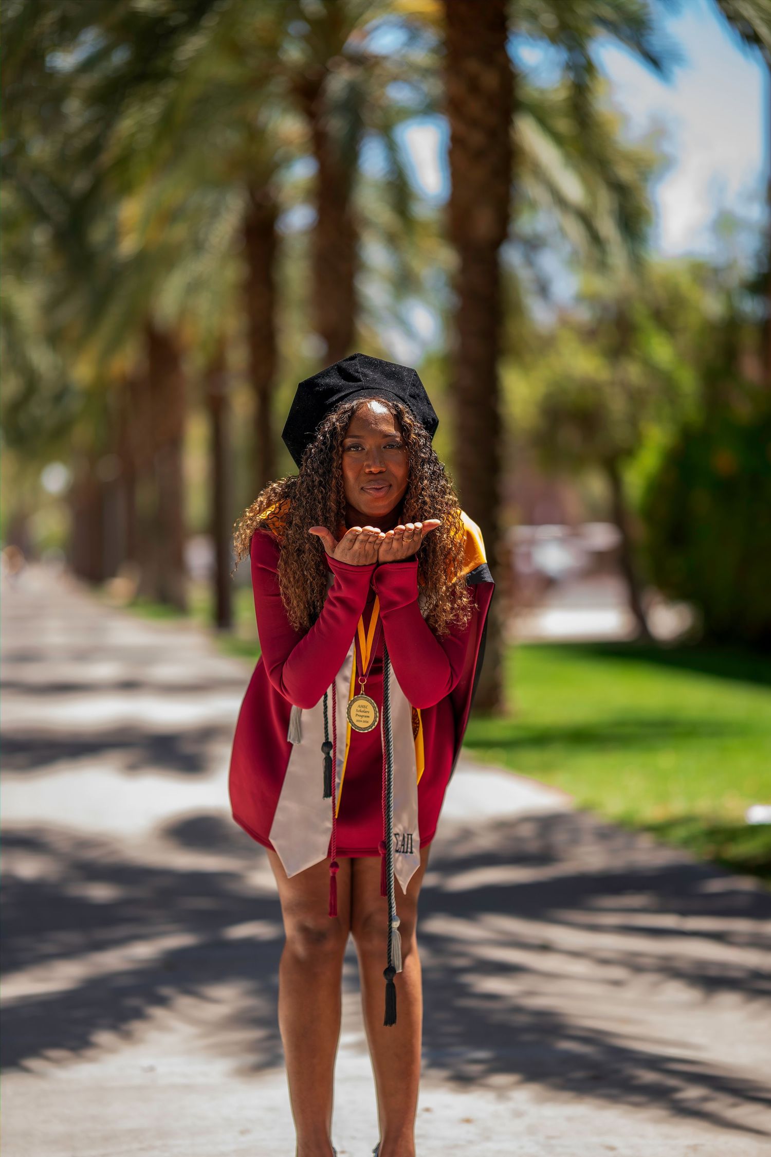 Joyful graduate in maroon gown and cap blows kiss on palm tree-lined campus walkway.