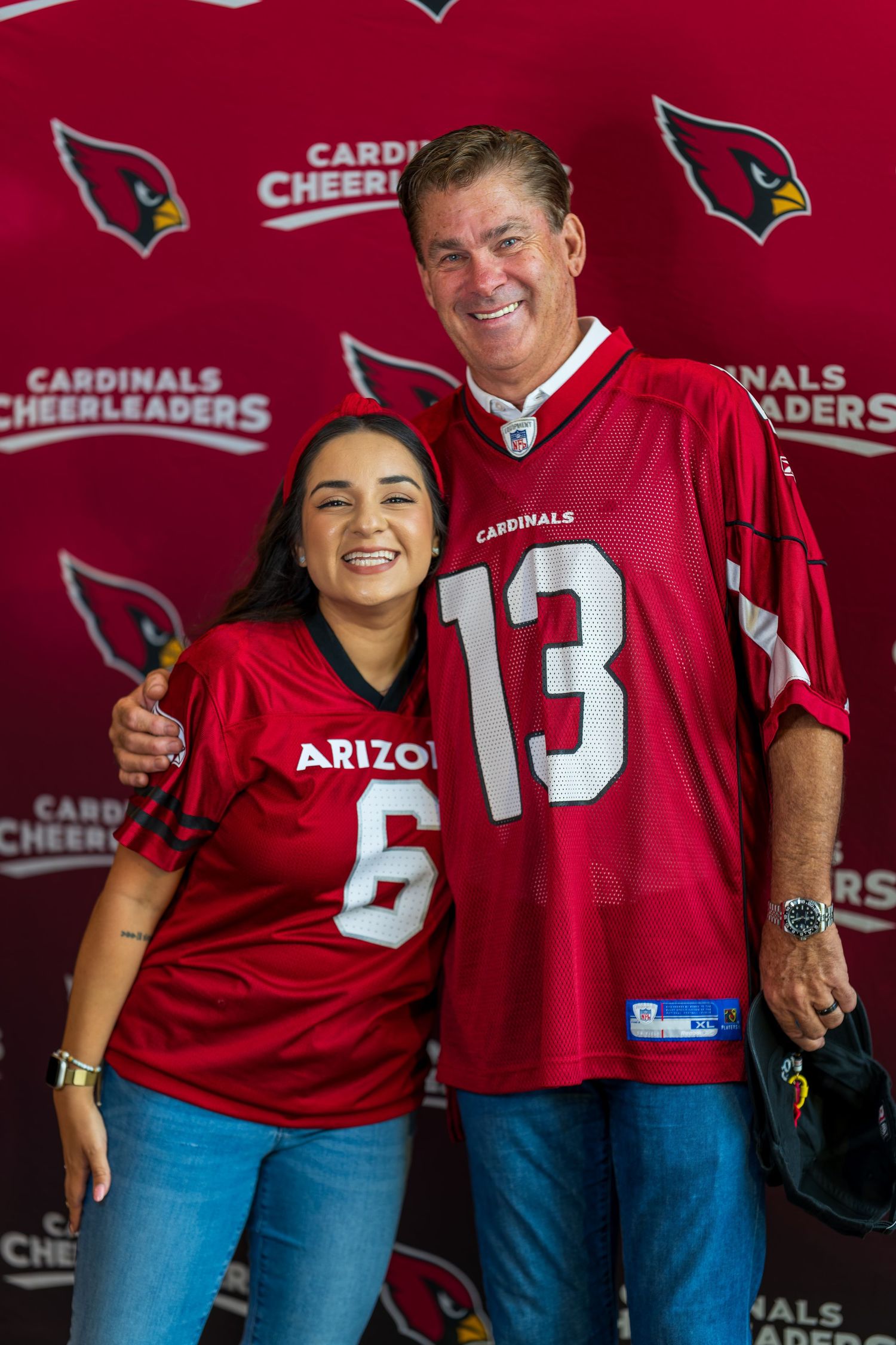 Sports fans wearing Arizona Cardinals jerseys pose against a red branded backdrop.