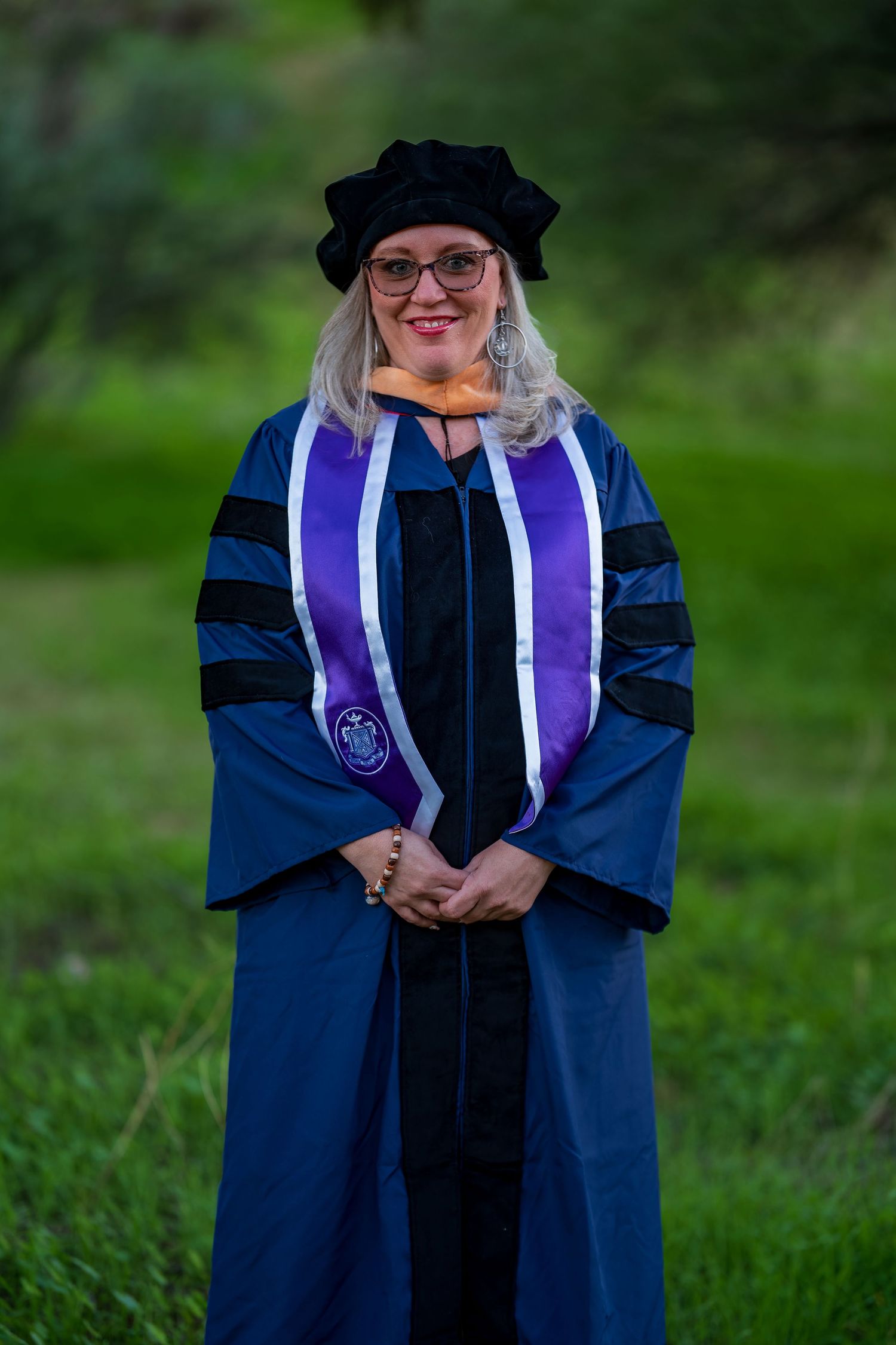 Graduate wearing doctoral regalia with blue and purple stoles stands on green grass.