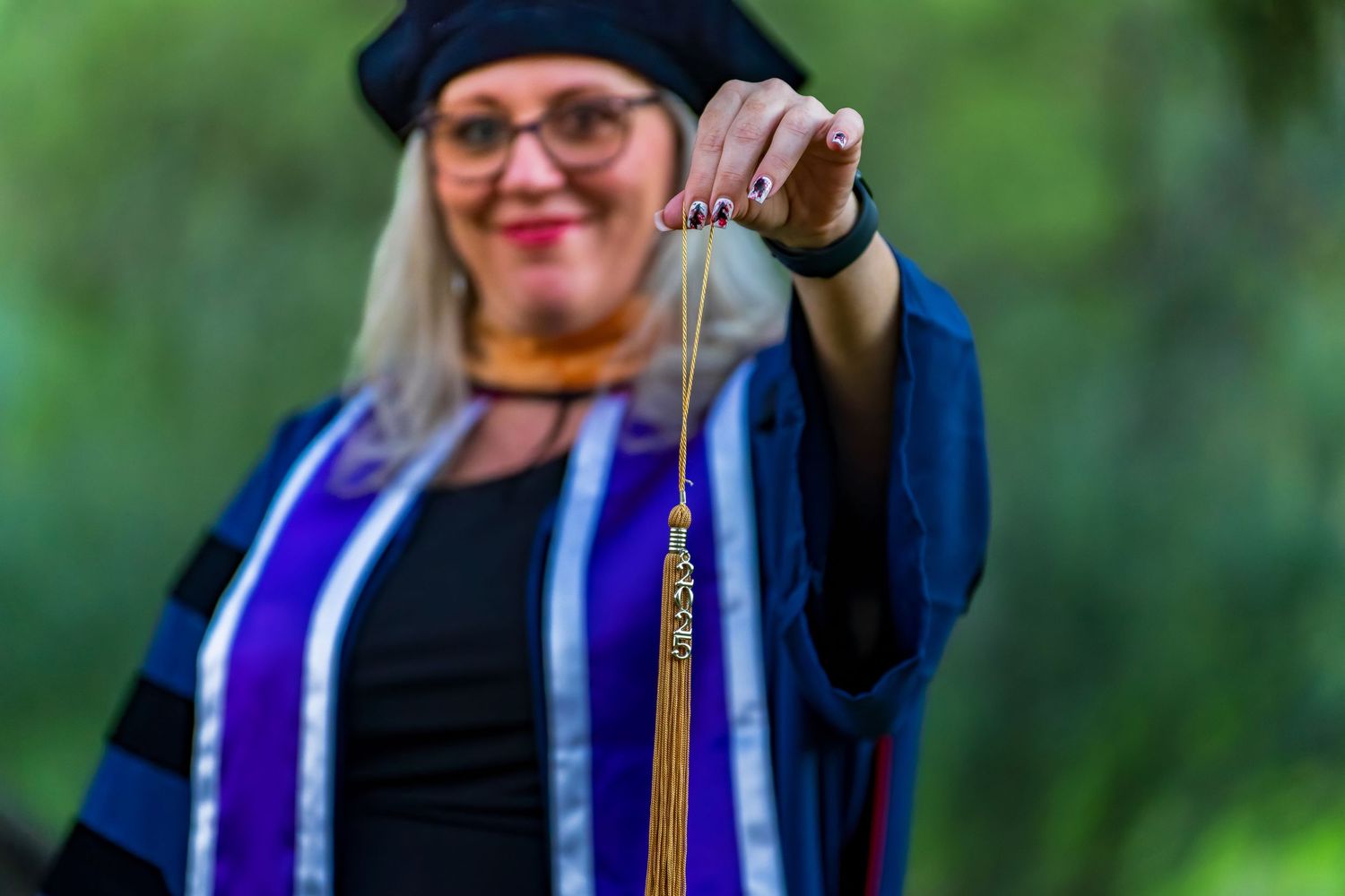 Graduate wearing cap and gown smiles while posing against blurred green background.